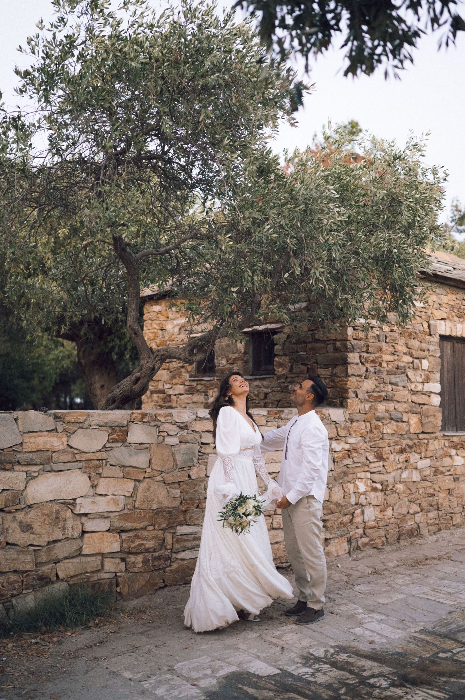 Bride and groom holding hands by rustic stone wall with olive tree, outdoor wedding portrait.