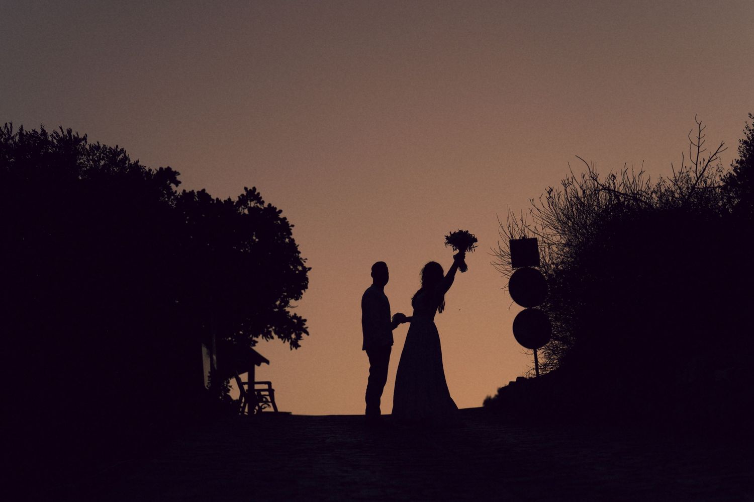 Silhouette of a couple at sunset, bride holding a bouquet, surrounded by trees in a romantic outdoor wedding scene.