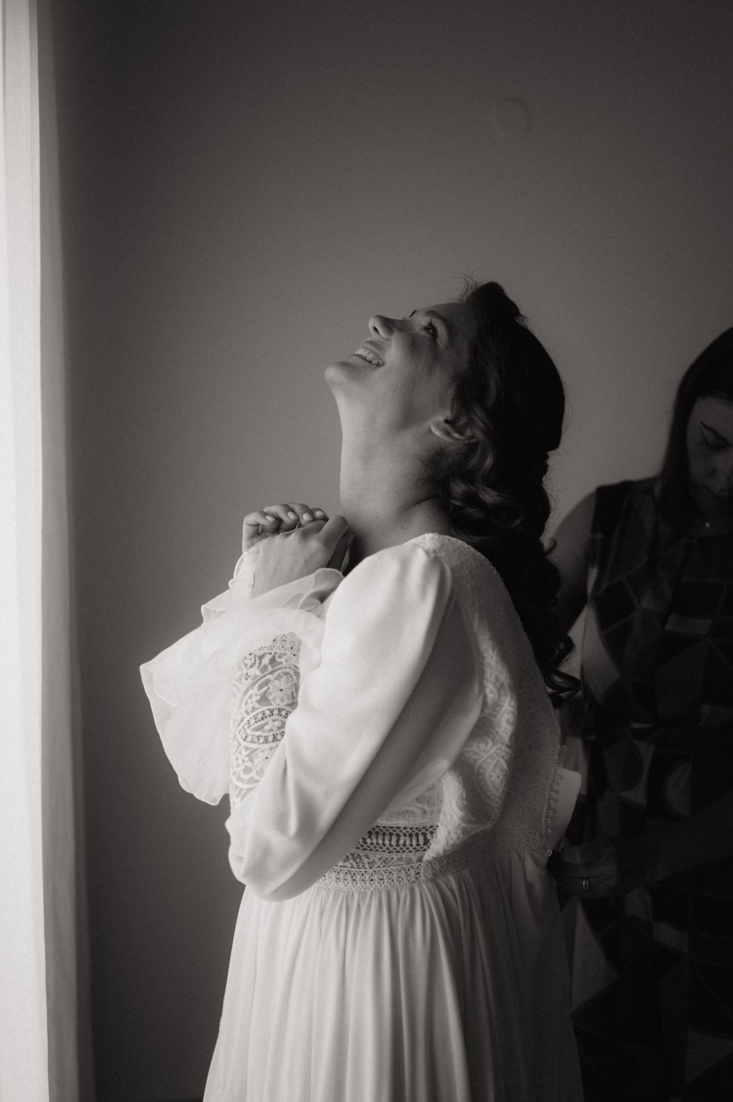 Black and white photo of a bride in a lace wedding dress laughing joyfully while getting ready.