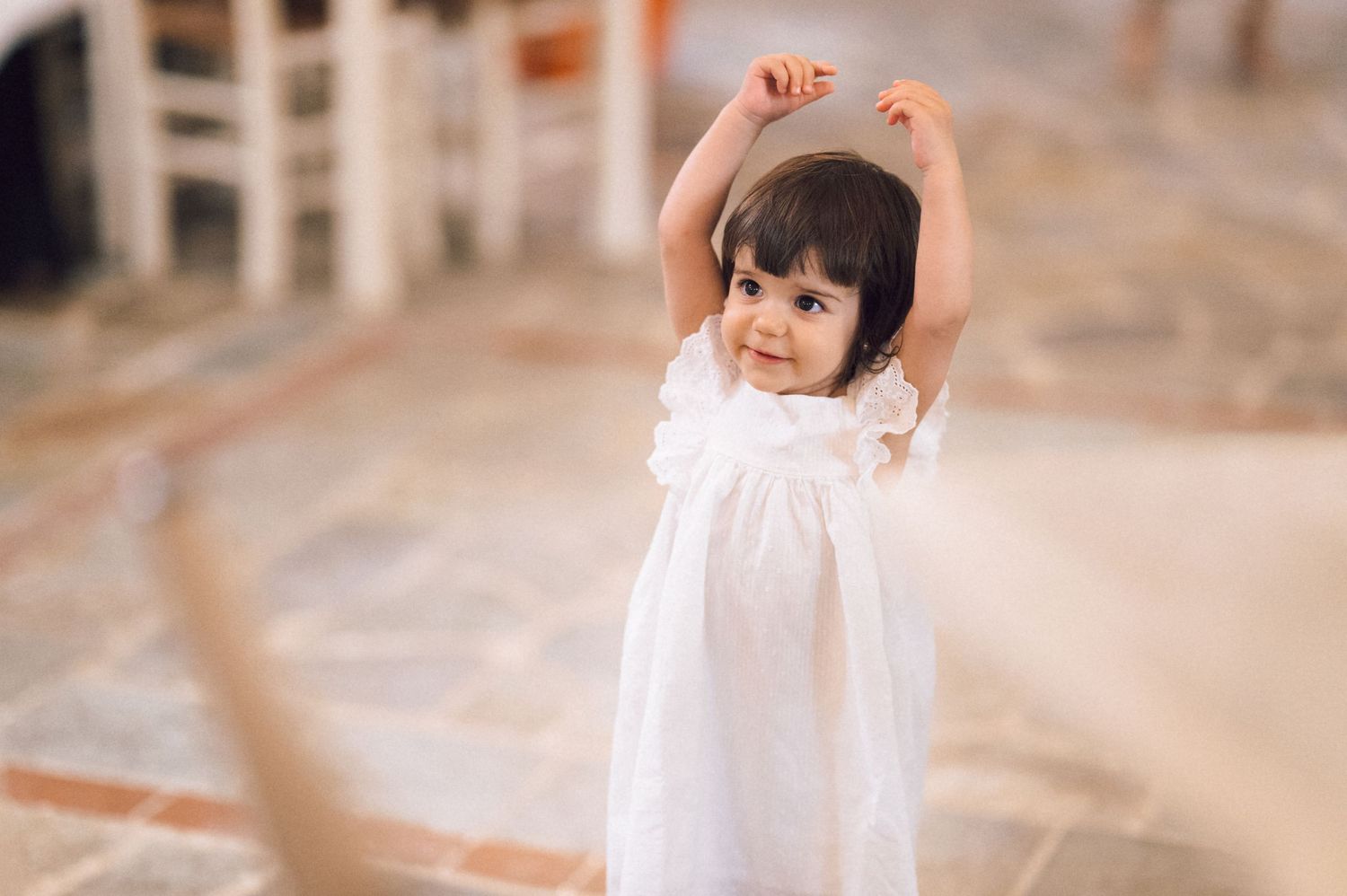 A joyful toddler girl in a white dress raises her arms while smiling indoors on a tiled floor.