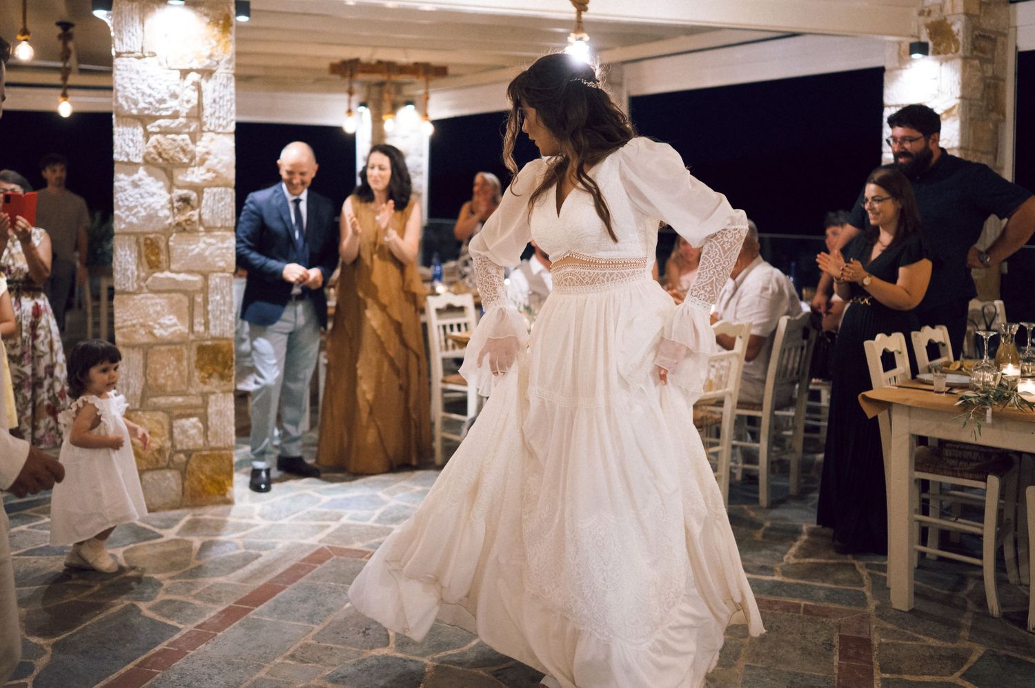 Bride in white gown dancing at wedding reception with guests clapping in rustic stone venue.