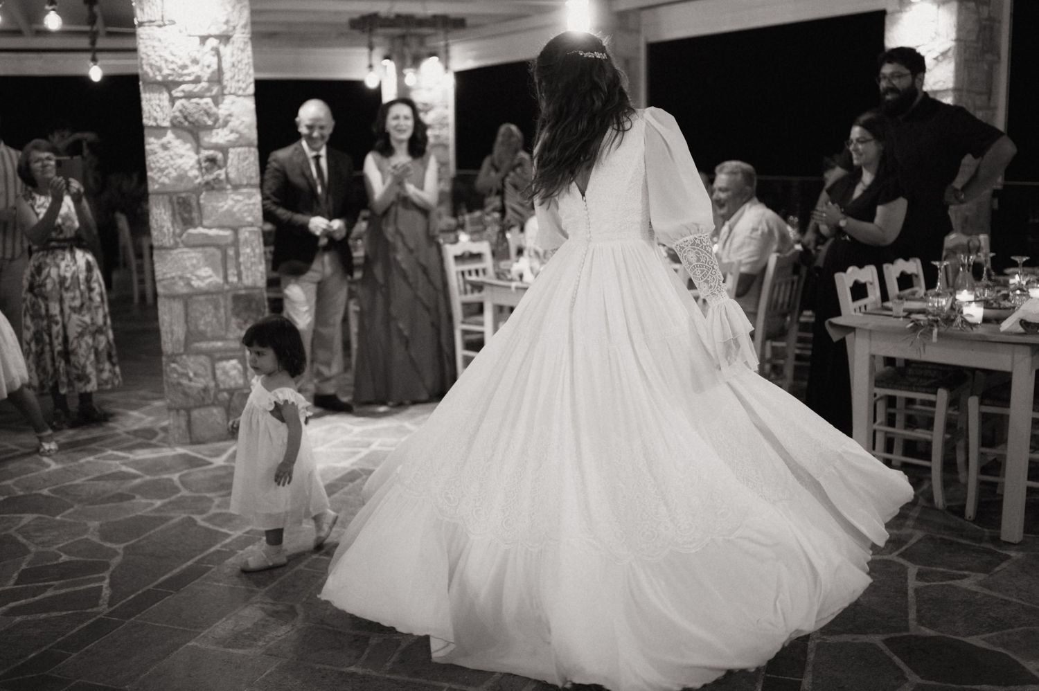 Bride twirling in white gown with flower girl at wedding reception, guests applauding in black and white photo.