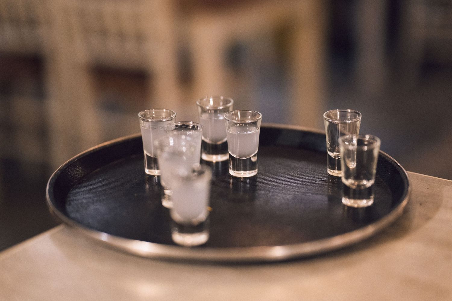 Shot glasses filled with clear liquor arranged on a round silver tray on a table.