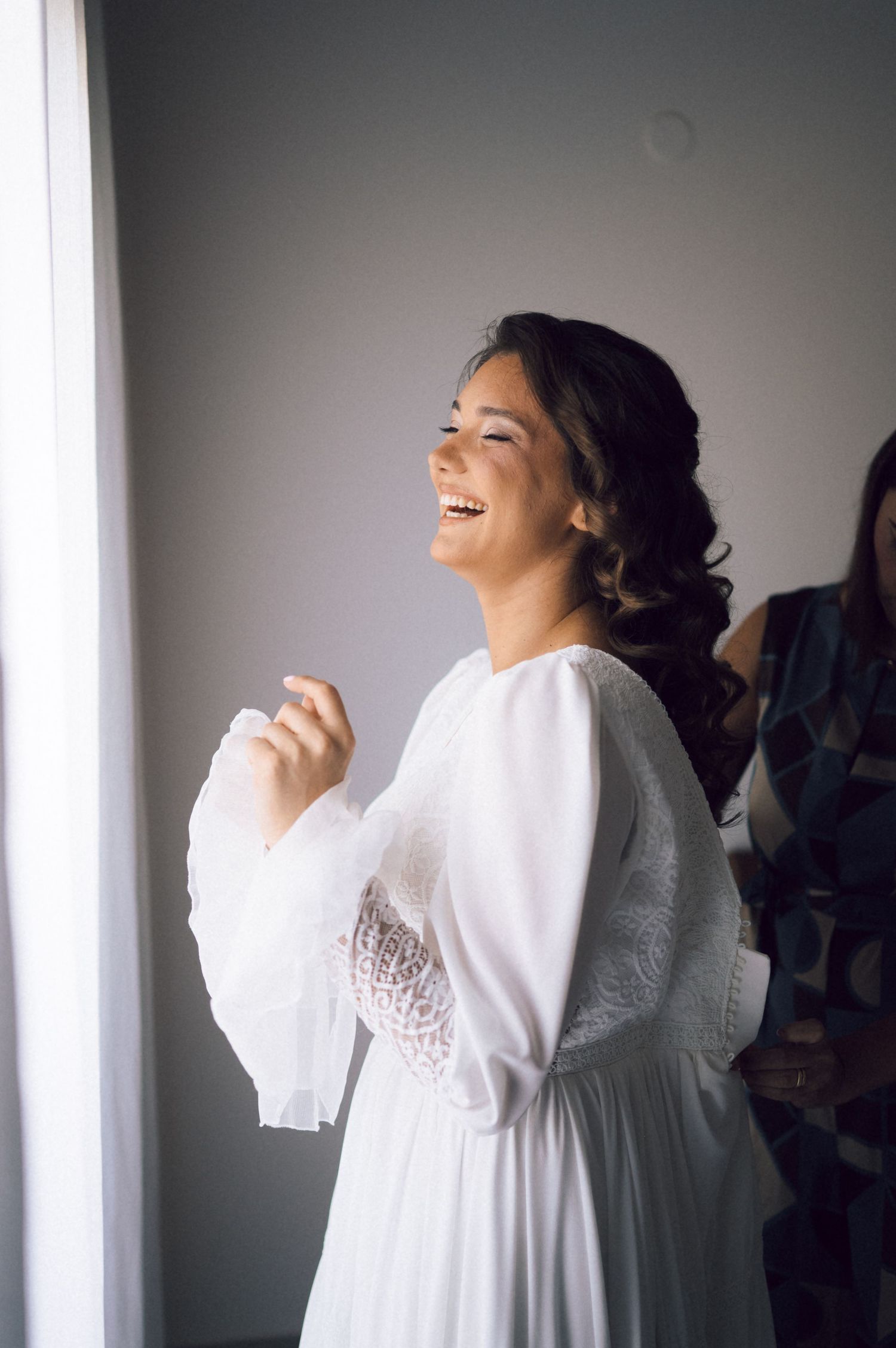 Joyful bride laughing in white lace wedding dress standing by bright window.