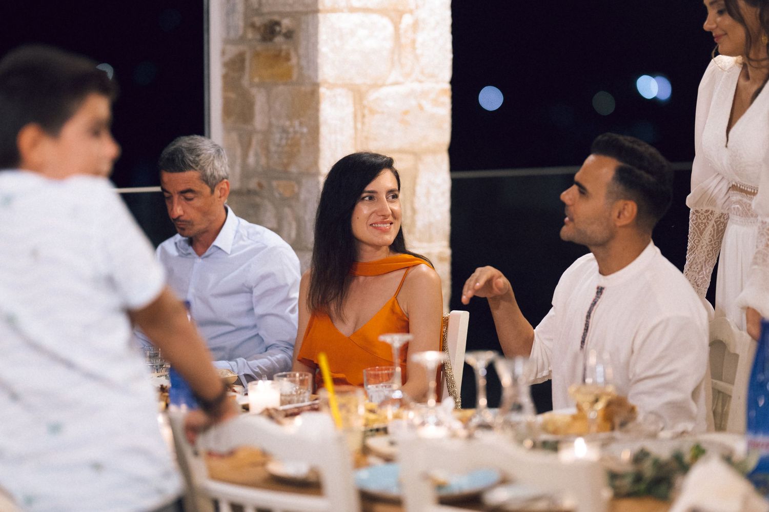 Family gathering around a candlelit dinner table at an outdoor evening event with stone walls in background.