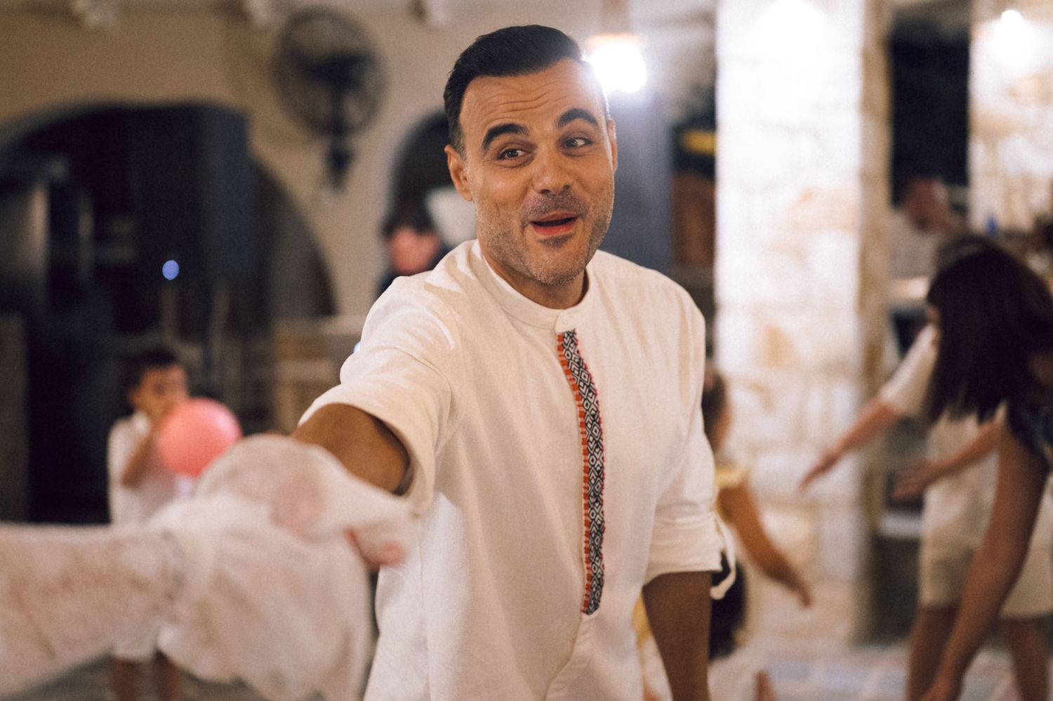 Man in white embroidered shirt smiling and reaching toward camera at an indoor celebration.