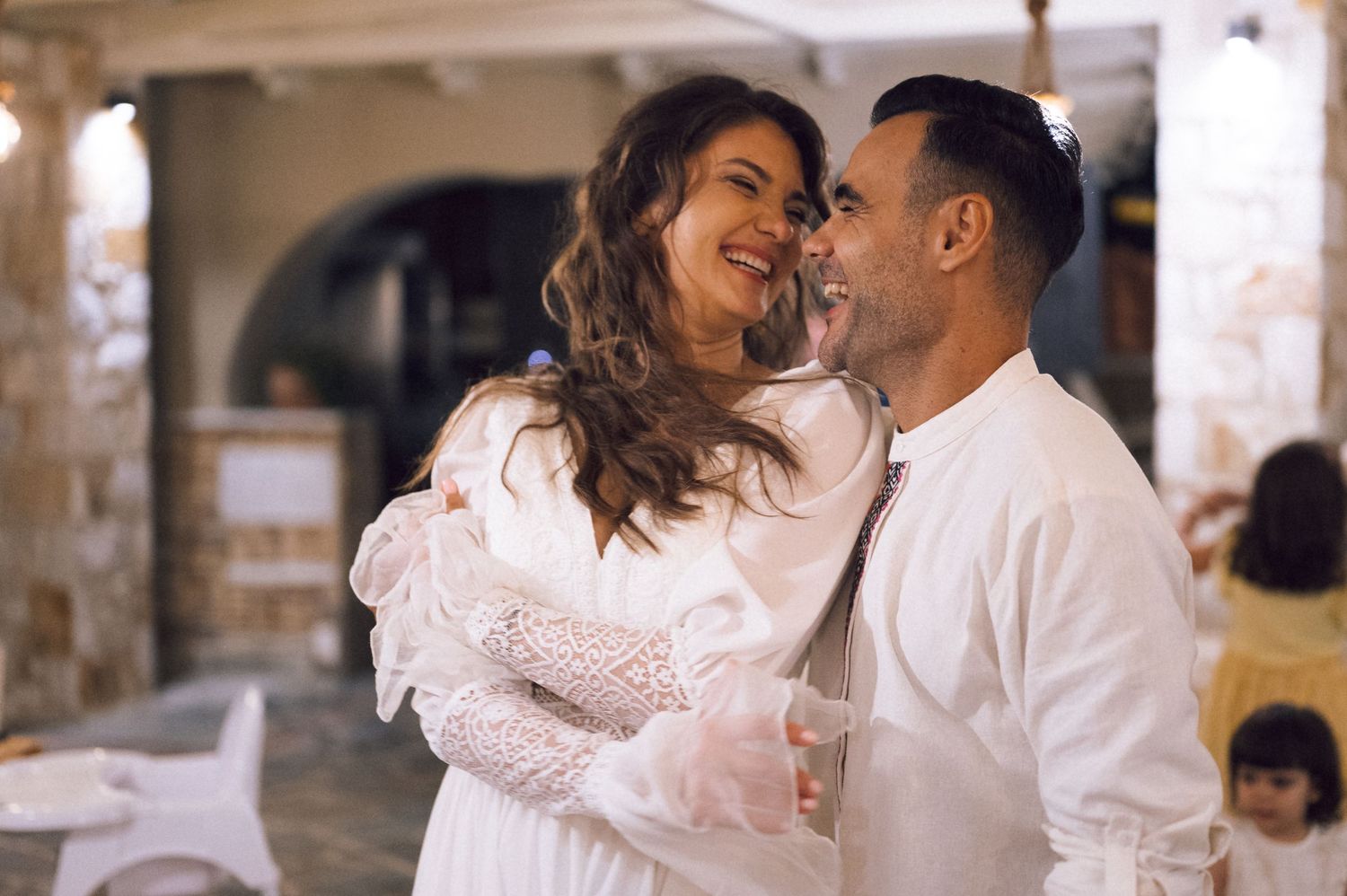 Couple in white outfits sharing a romantic moment, smiling at each other at an outdoor Mediterranean venue.