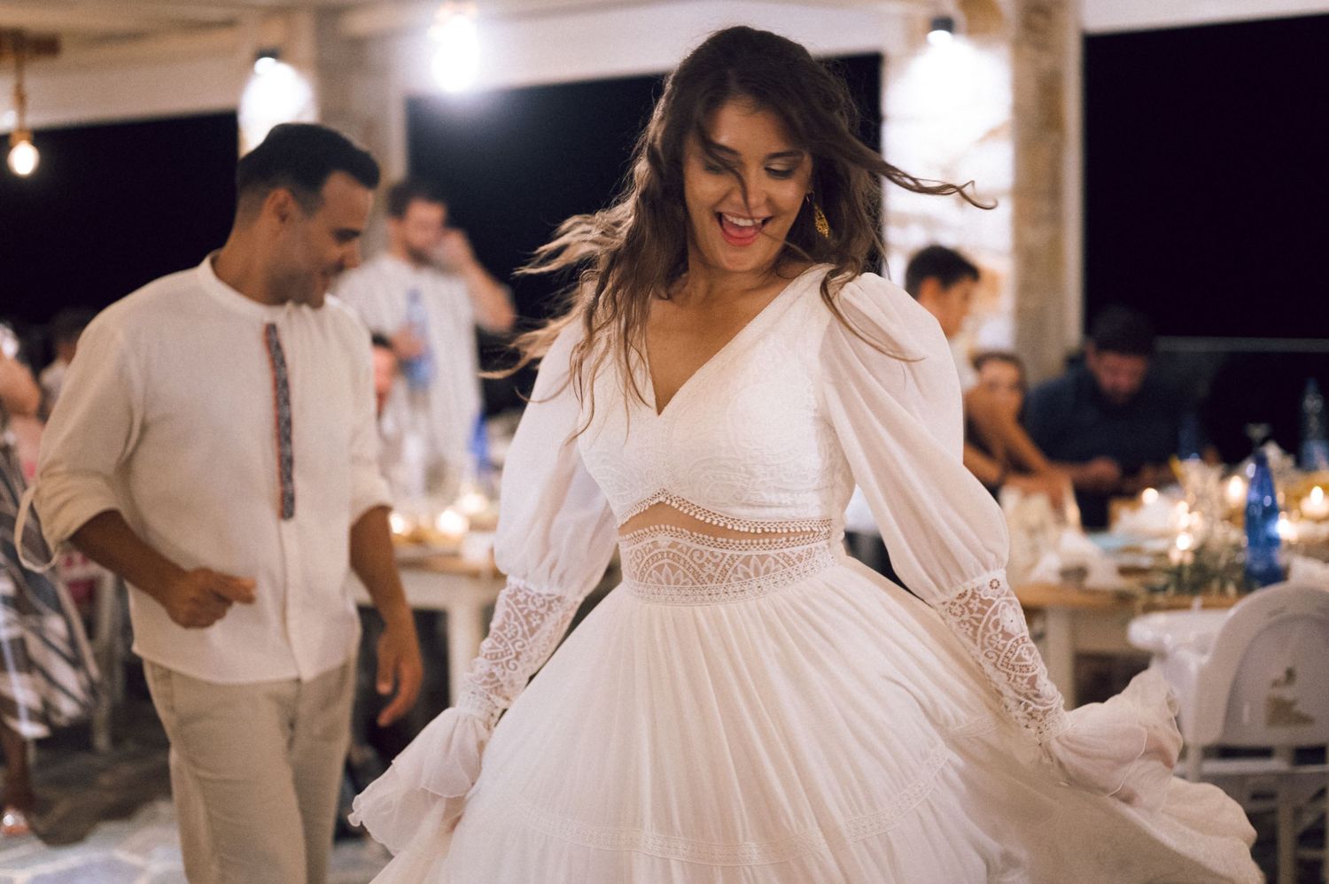 Woman in white dress dancing joyfully at an outdoor evening wedding reception with guests in background.
