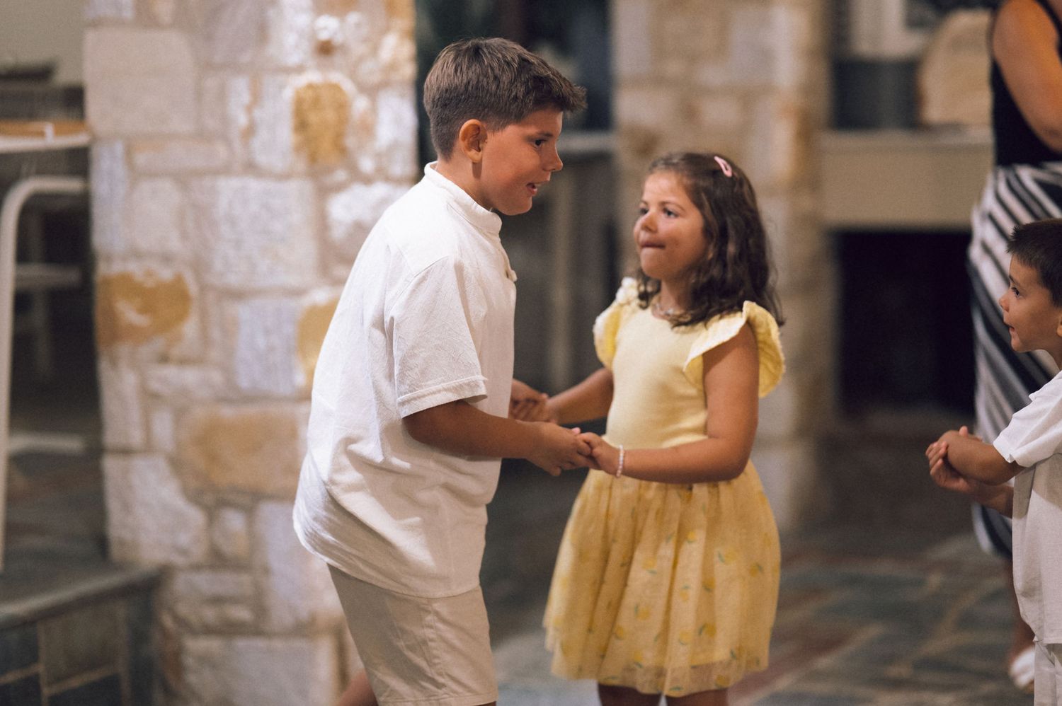 Two children, a boy in white and a girl in yellow dress, interact outdoors near stone architecture.