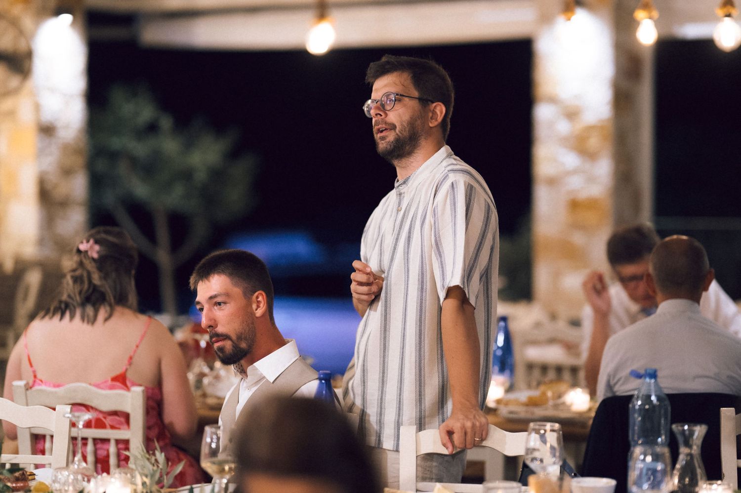 Man standing and speaking at an outdoor wedding reception dinner with guests seated at decorated tables.