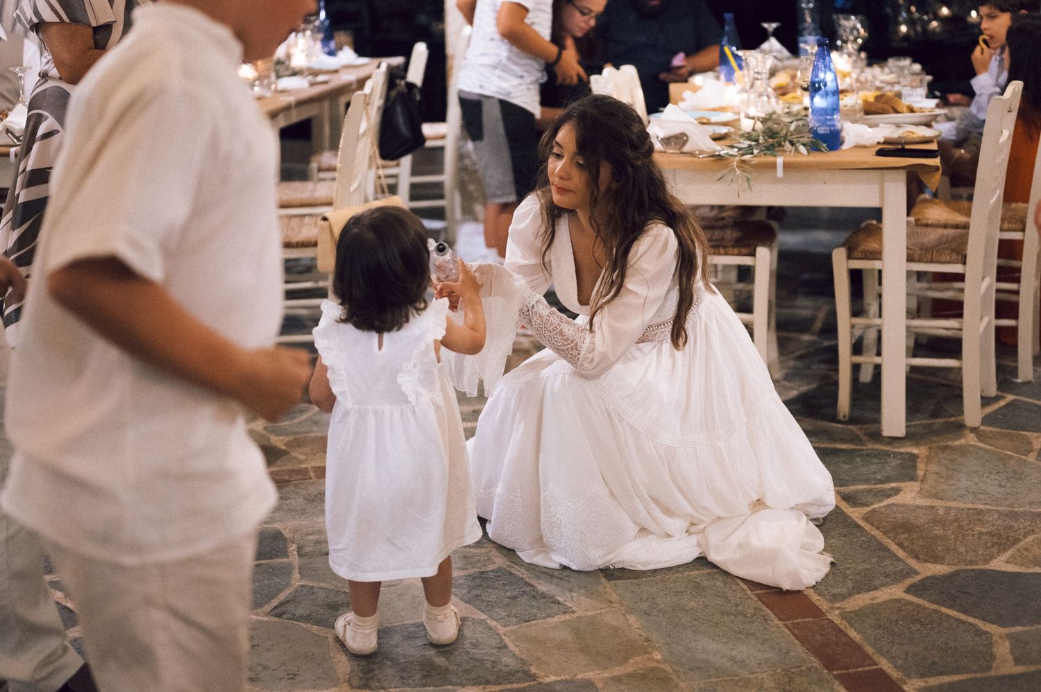 Bride in white gown crouches to interact with young girl in white dress at an outdoor wedding reception.