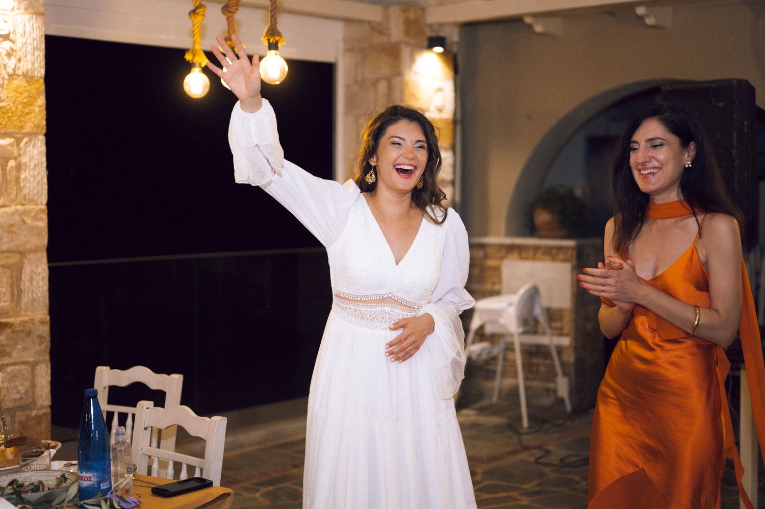 Pregnant bride in white dress laughing and waving at wedding reception, with guest in orange dress clapping.