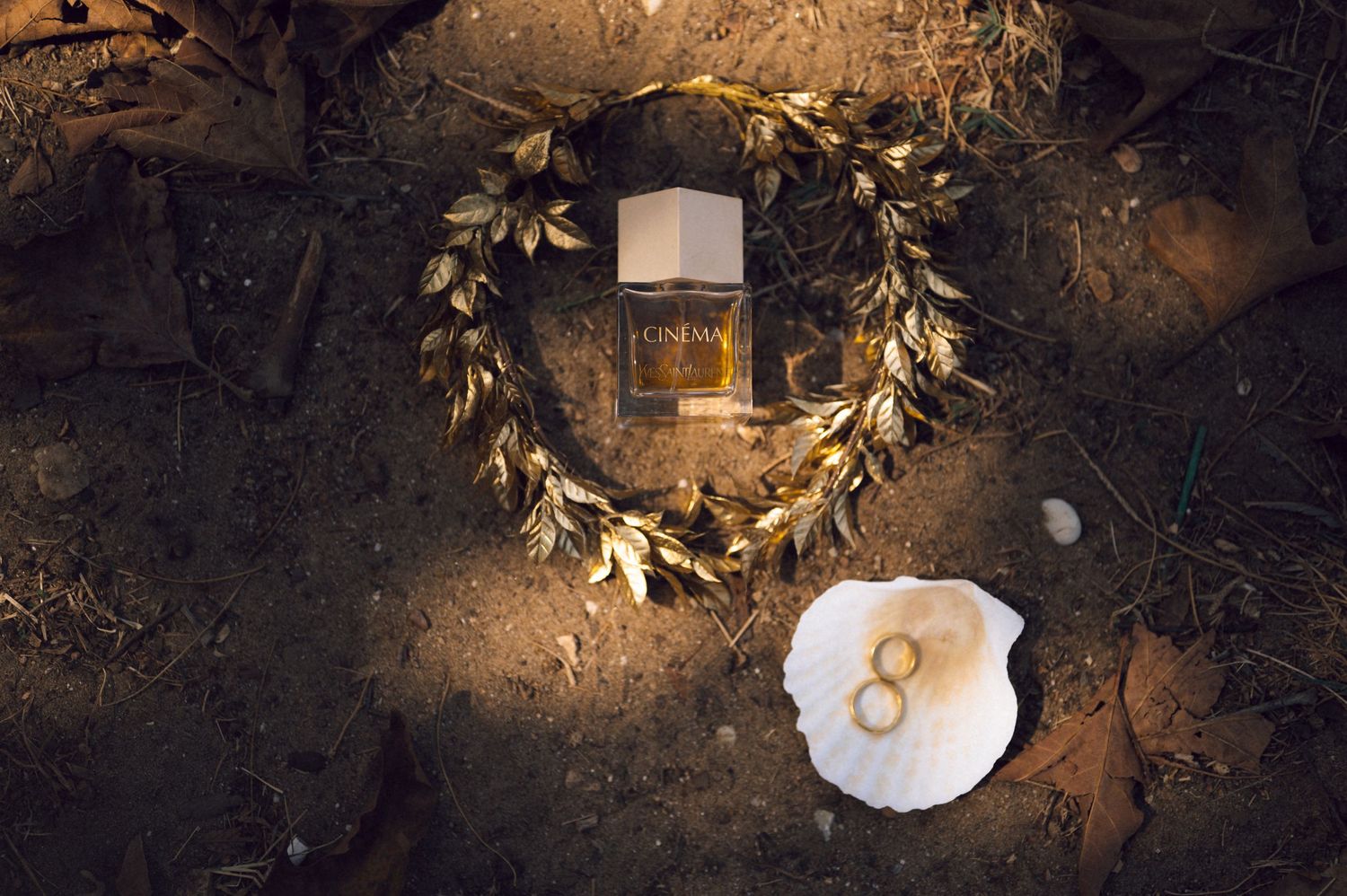 Gold leaf heart wreath with perfume bottle and wedding rings on seashell, arranged on rustic ground.