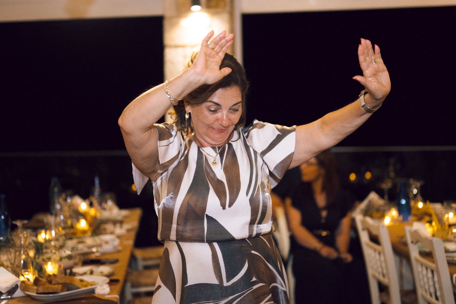 Woman in patterned dress dancing with raised hands at an elegant outdoor dinner event at night.