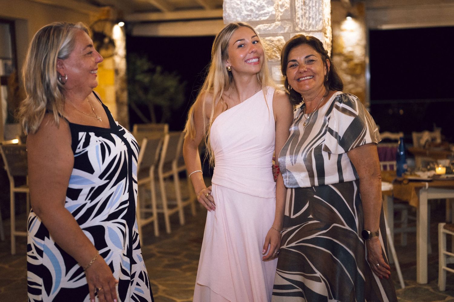 Three women smiling and posing together at an elegant outdoor evening event, with the center woman in a pink dress.