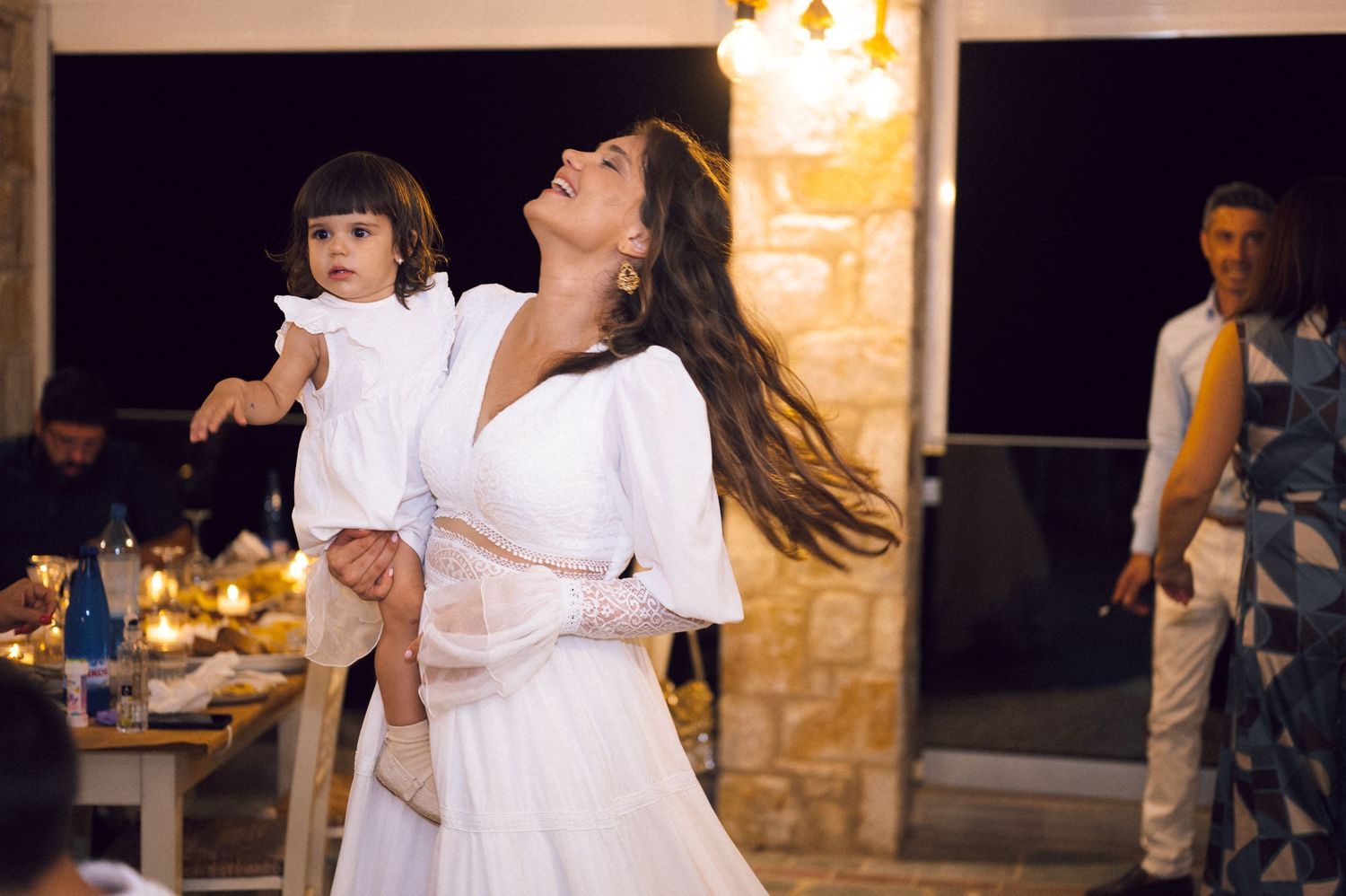 Woman in white dress dancing while holding a toddler girl at an elegant outdoor evening celebration.