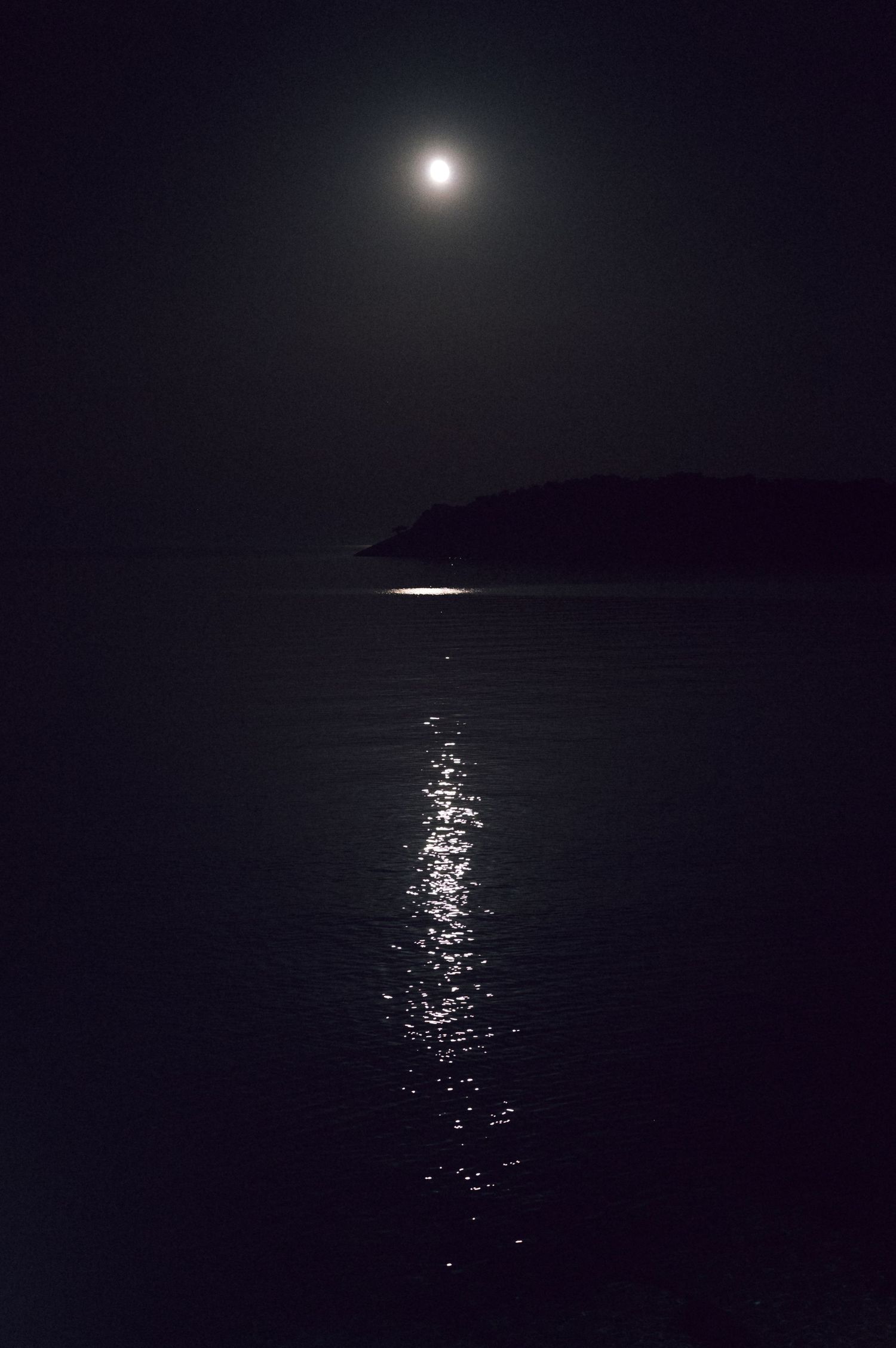 Moonlit night seascape with glowing reflection on calm ocean water and dark silhouetted island in background.