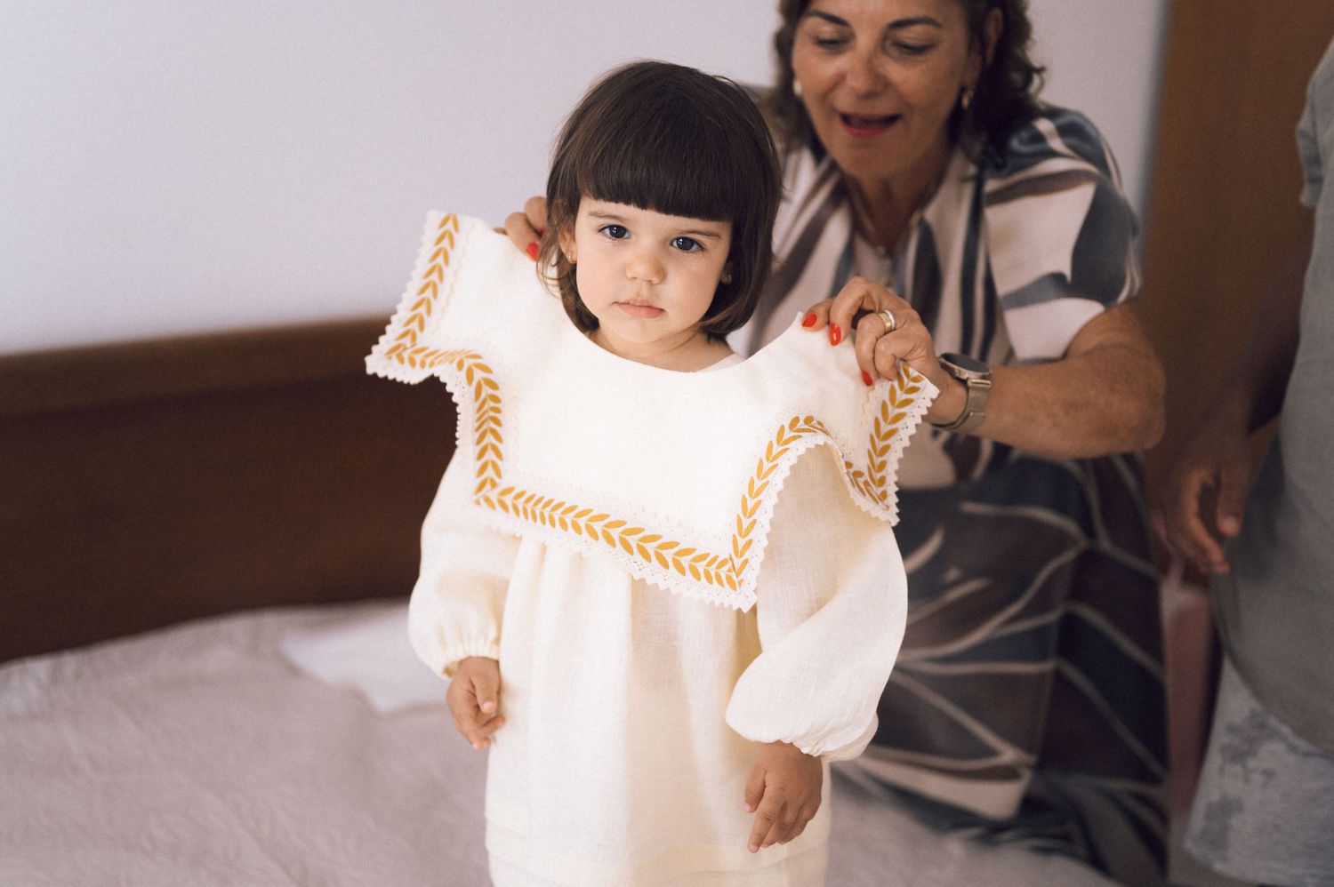 A grandmother helps a toddler girl try on a white embroidered dress with gold trim while standing on a bed.