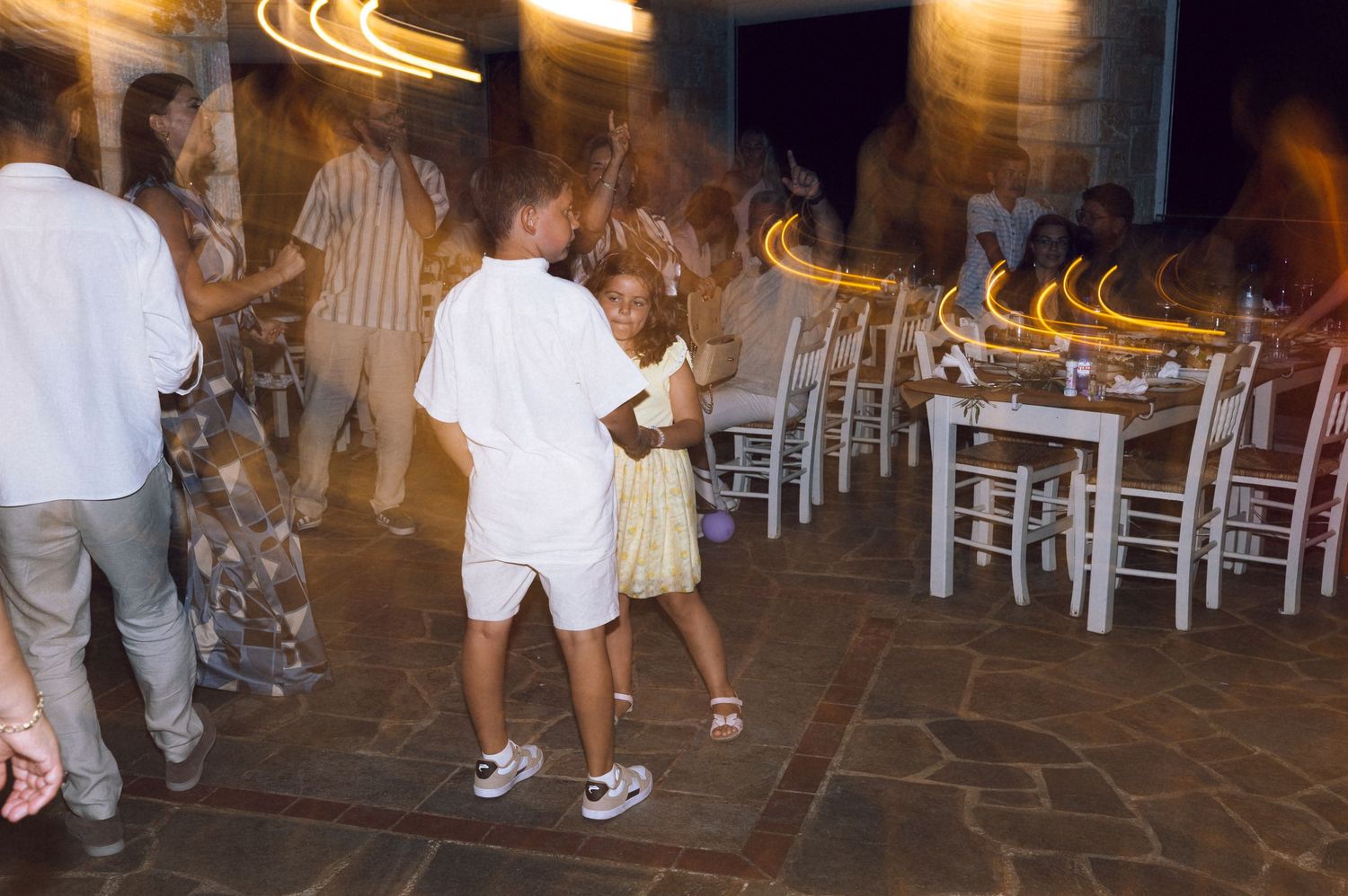 Two children dancing together at an outdoor evening party with white chairs and tables in the background.