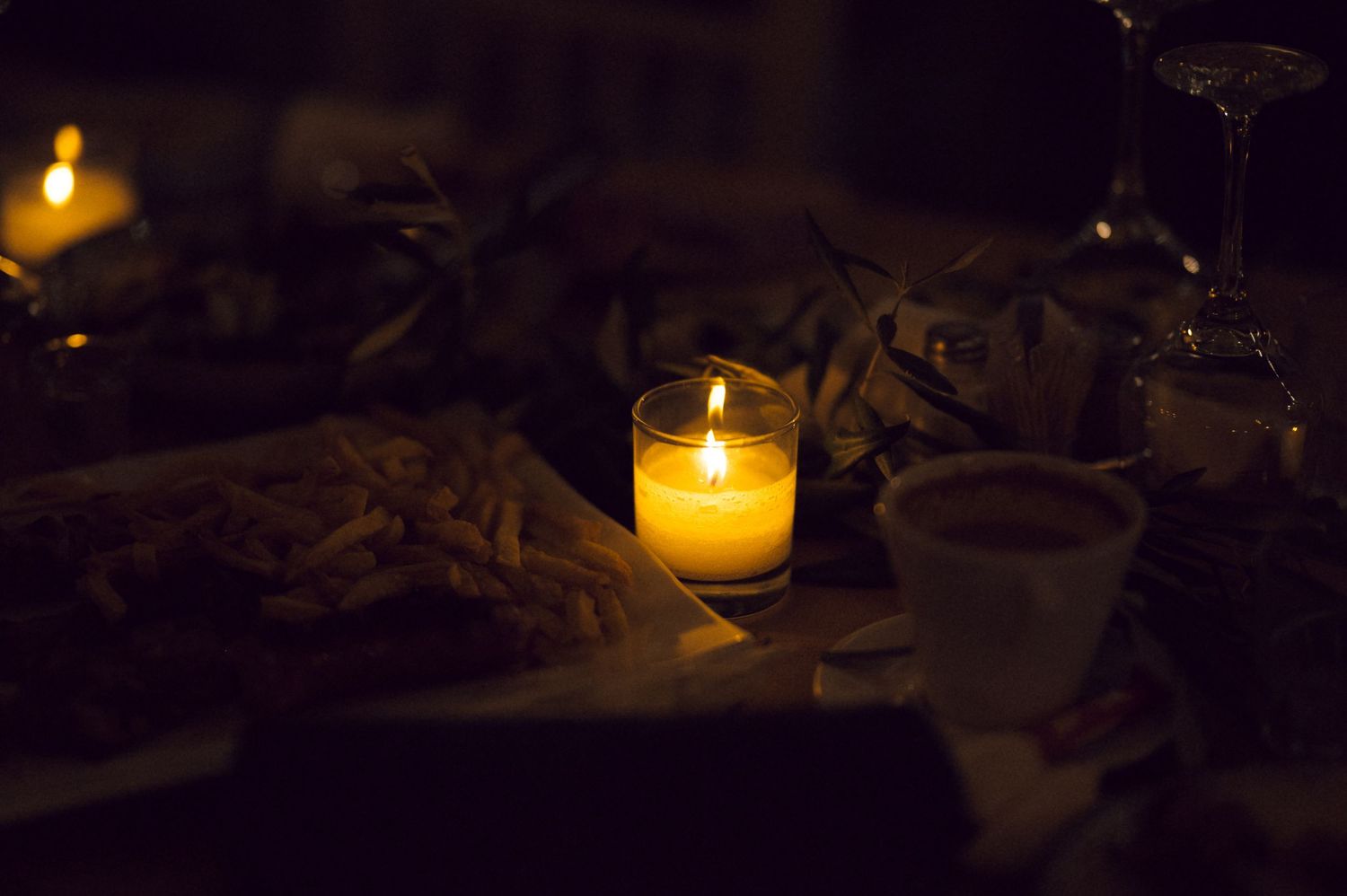 Romantic candlelit dinner table with glowing candle, wine glasses, and food in dark ambiance.