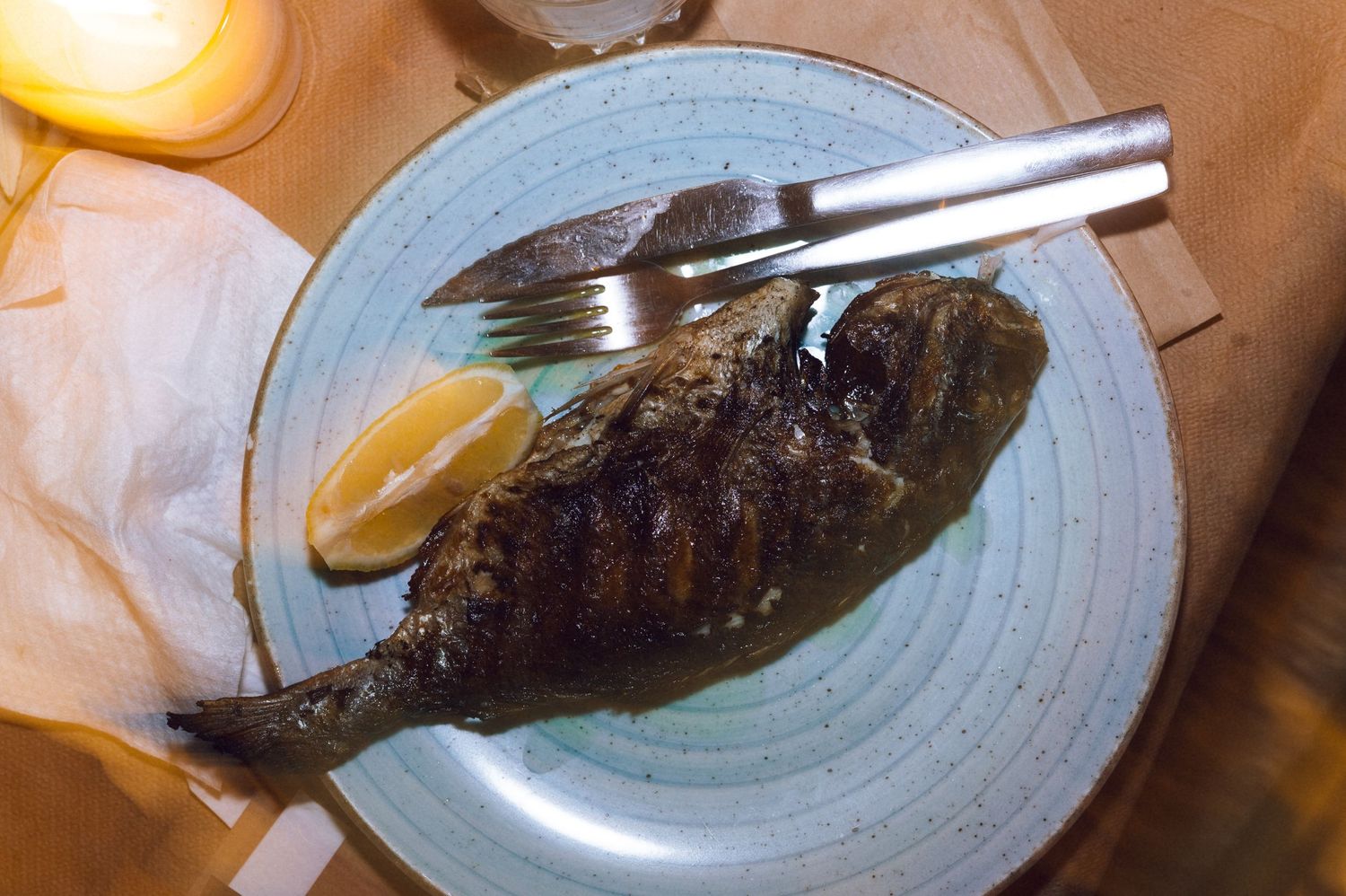 Grilled whole fish served on a blue plate with lemon wedges and cutlery on a rustic table.