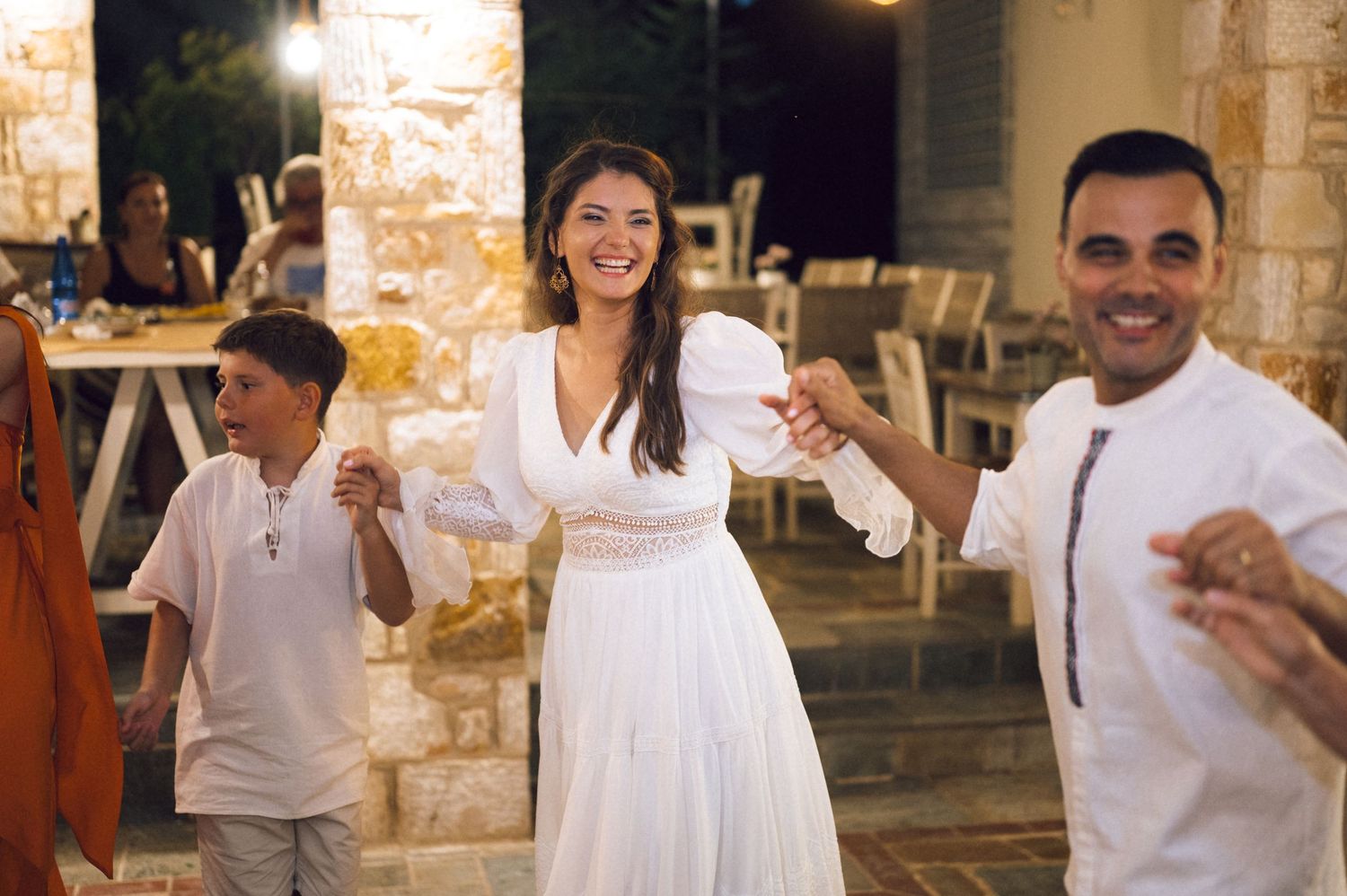 Woman in white dress dancing joyfully with man and child at an outdoor evening celebration.