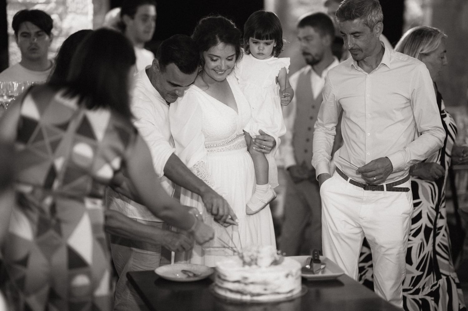 Black and white photo of a pregnant woman and man cutting a cake surrounded by family at a baby shower celebration.