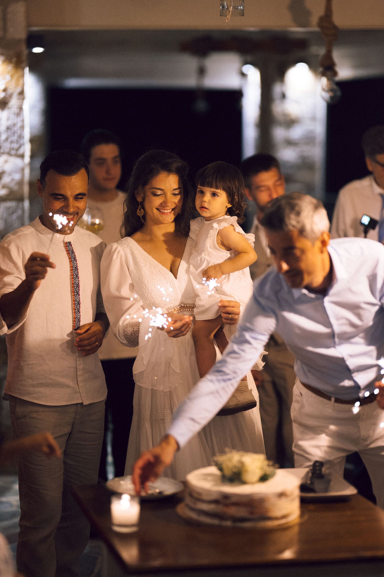 Wedding couple with sparklers cutting cake, bride holding child in white dress at elegant evening reception.