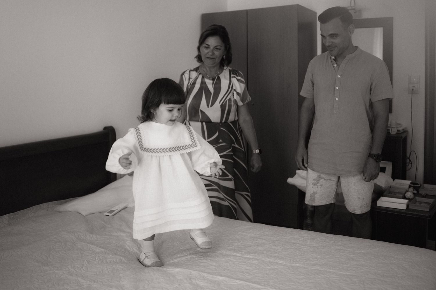 A young girl in a white dress stands on a bed while her parents watch in a black and white family photo.