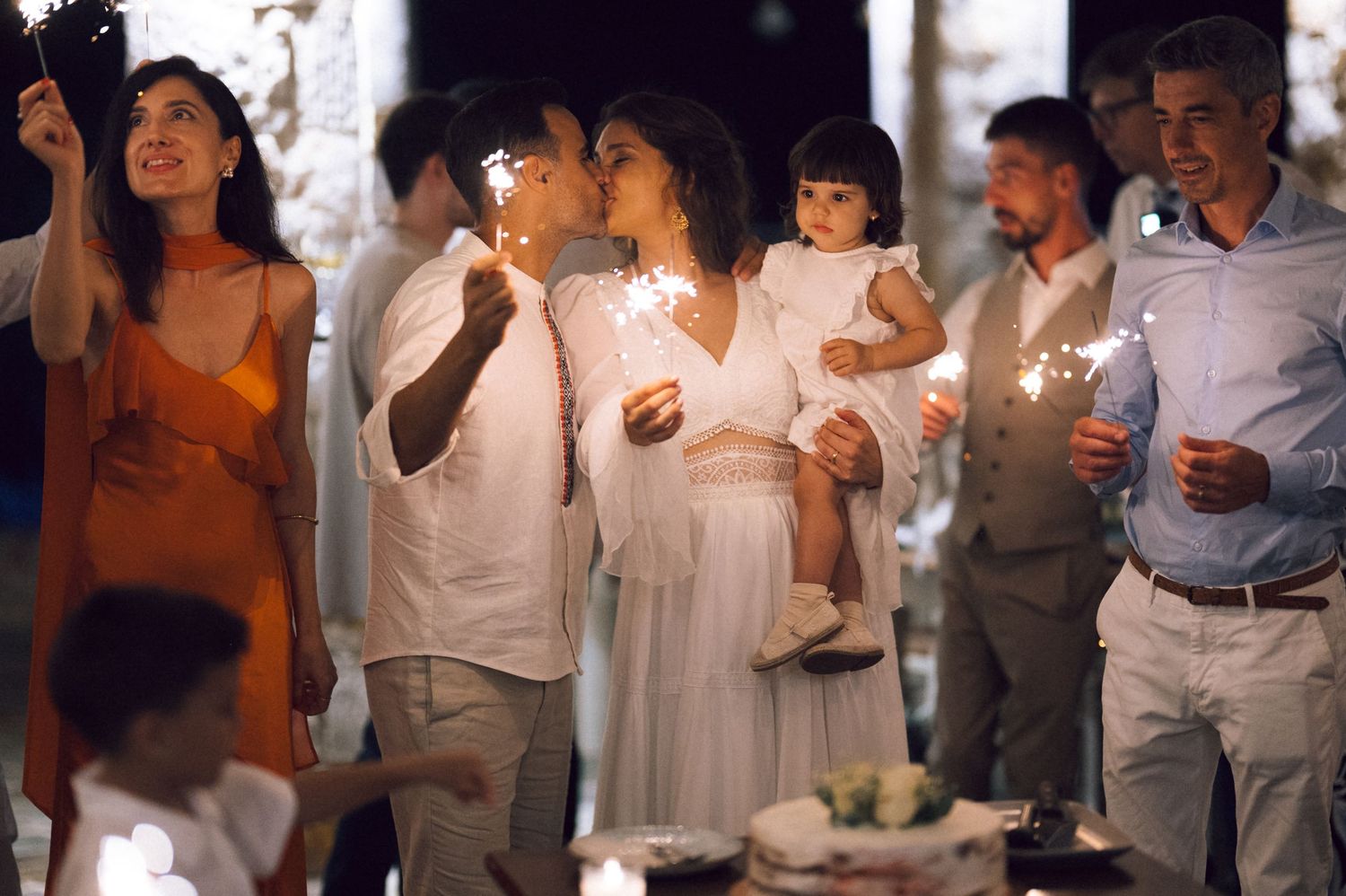 Guests celebrating at an outdoor party holding sparklers, with a couple kissing while holding a child in a festive setting.