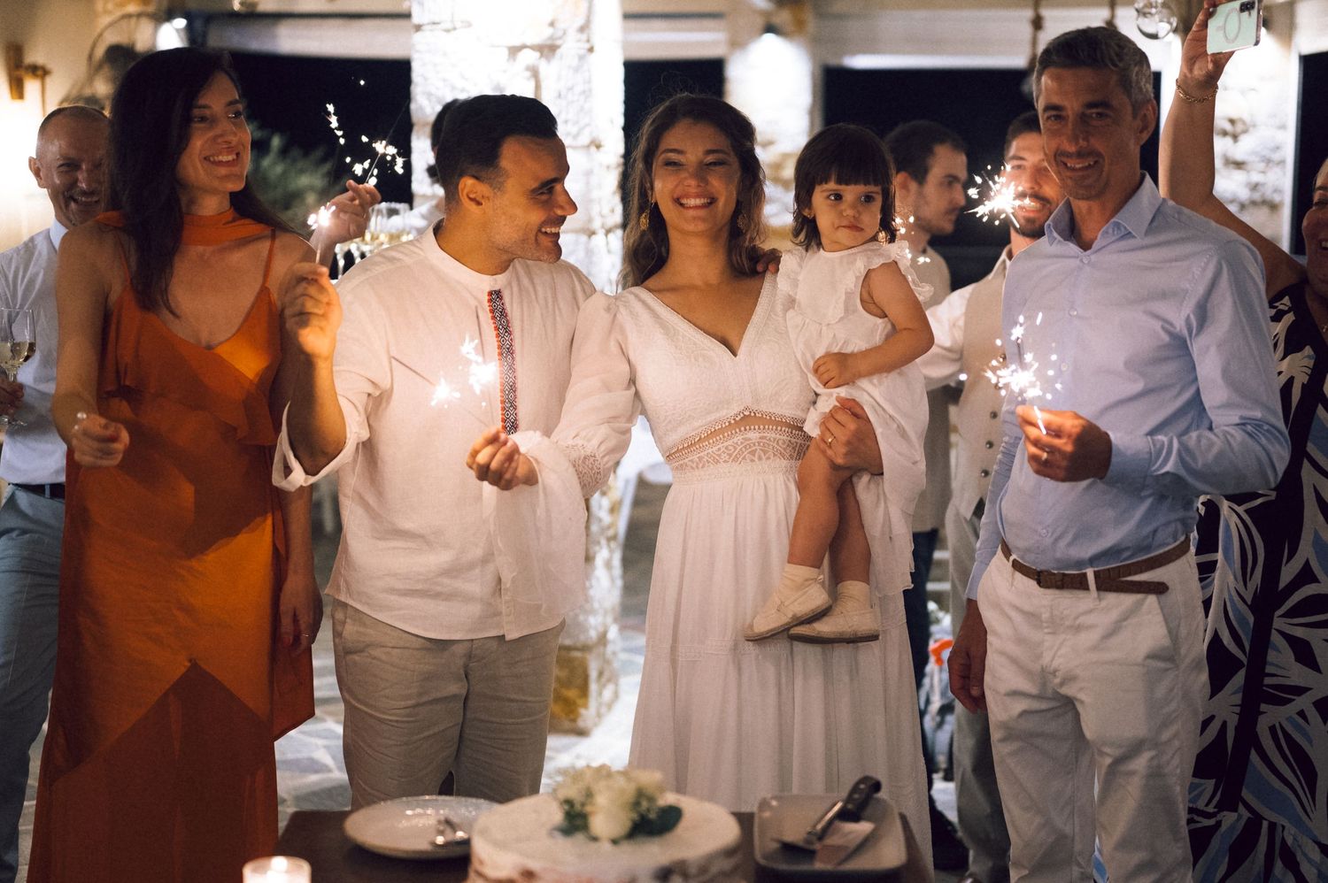Group of people celebrating with sparklers around a white wedding cake at an elegant outdoor evening reception.