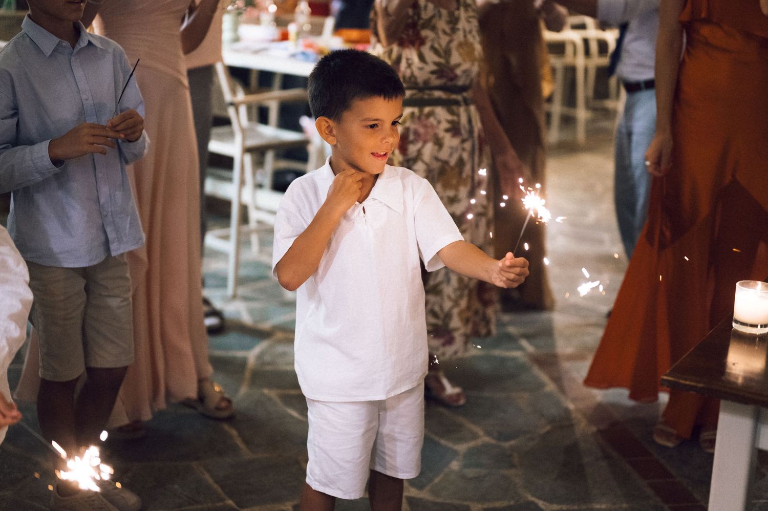 Young boy in white outfit holding sparklers at a festive outdoor celebration event.