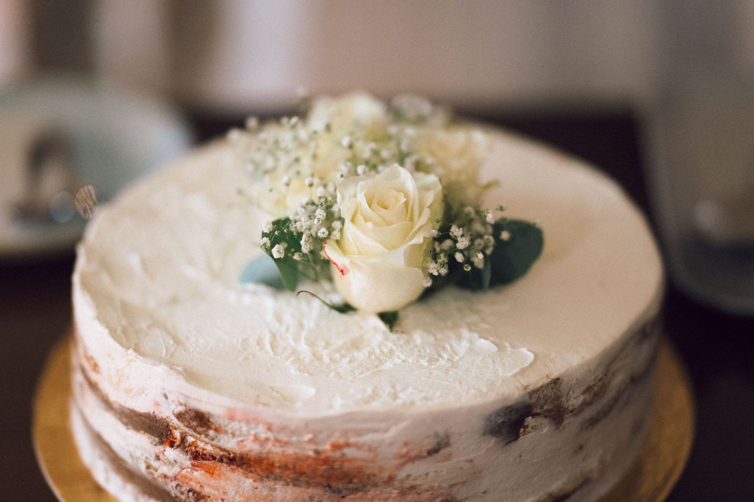 Elegant naked wedding cake with white frosting, topped with a white rose and baby's breath flowers on a gold stand.