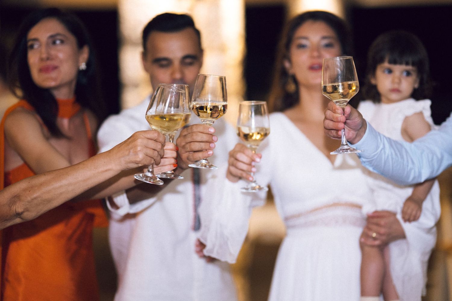 Group of adults toasting with white wine glasses at a celebration, woman in white dress holding a child.