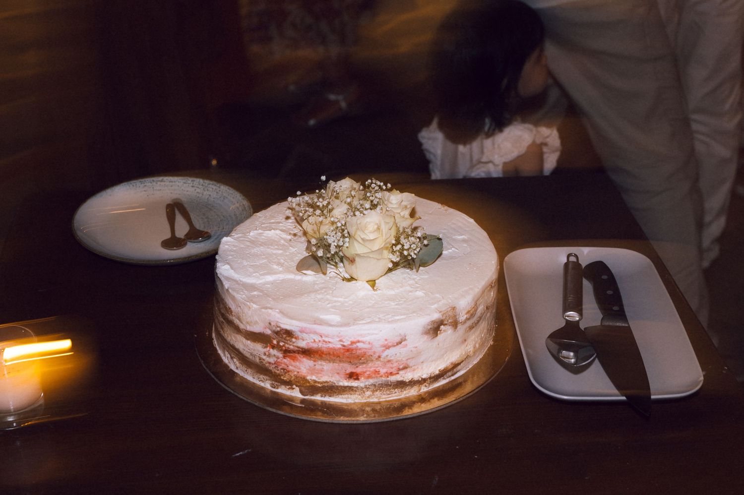 Elegant naked wedding cake topped with white roses and baby's breath on a dark wooden table.
