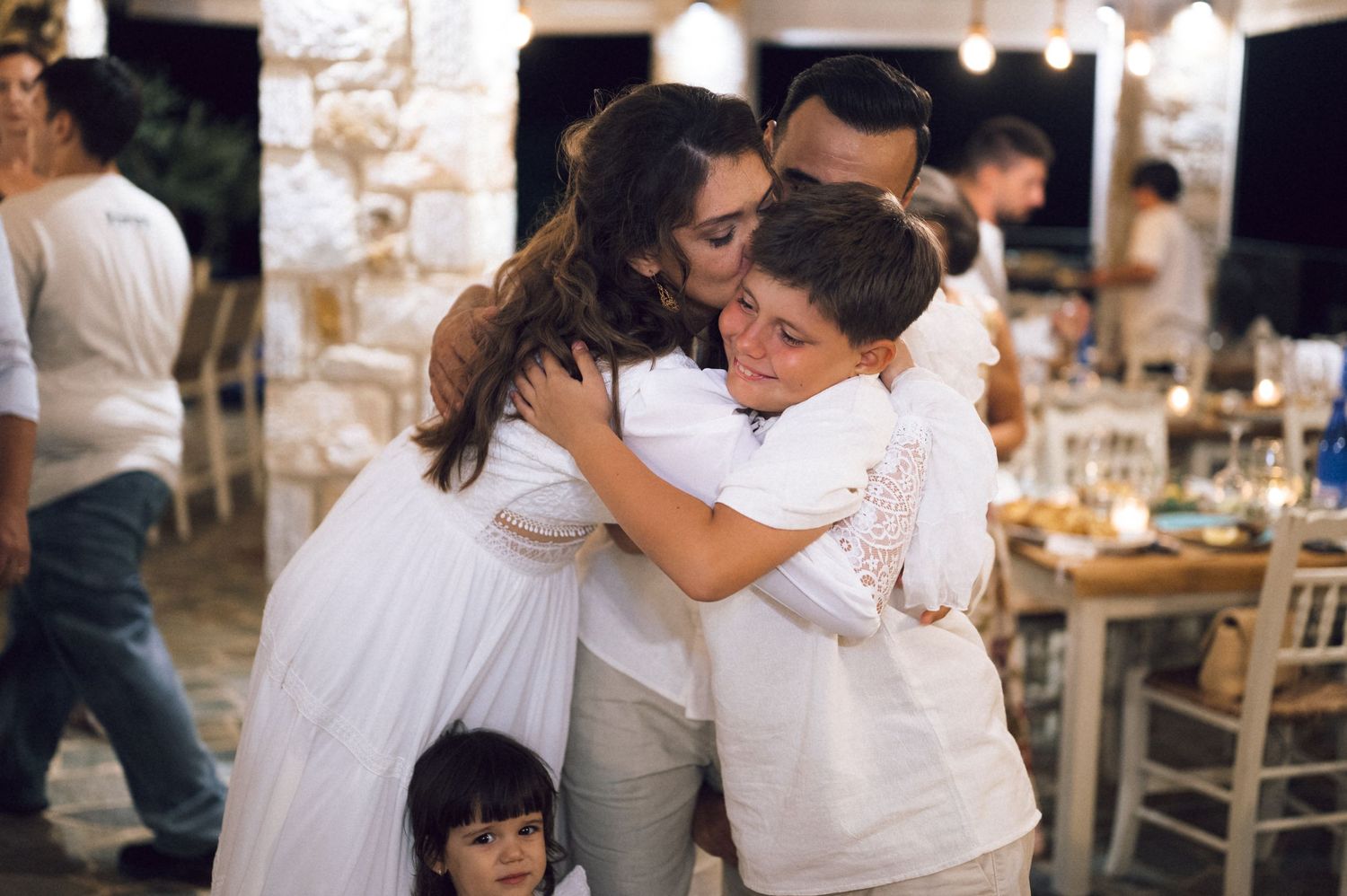 Family sharing a warm group hug at an outdoor celebration, dressed in white, with a young child nearby.