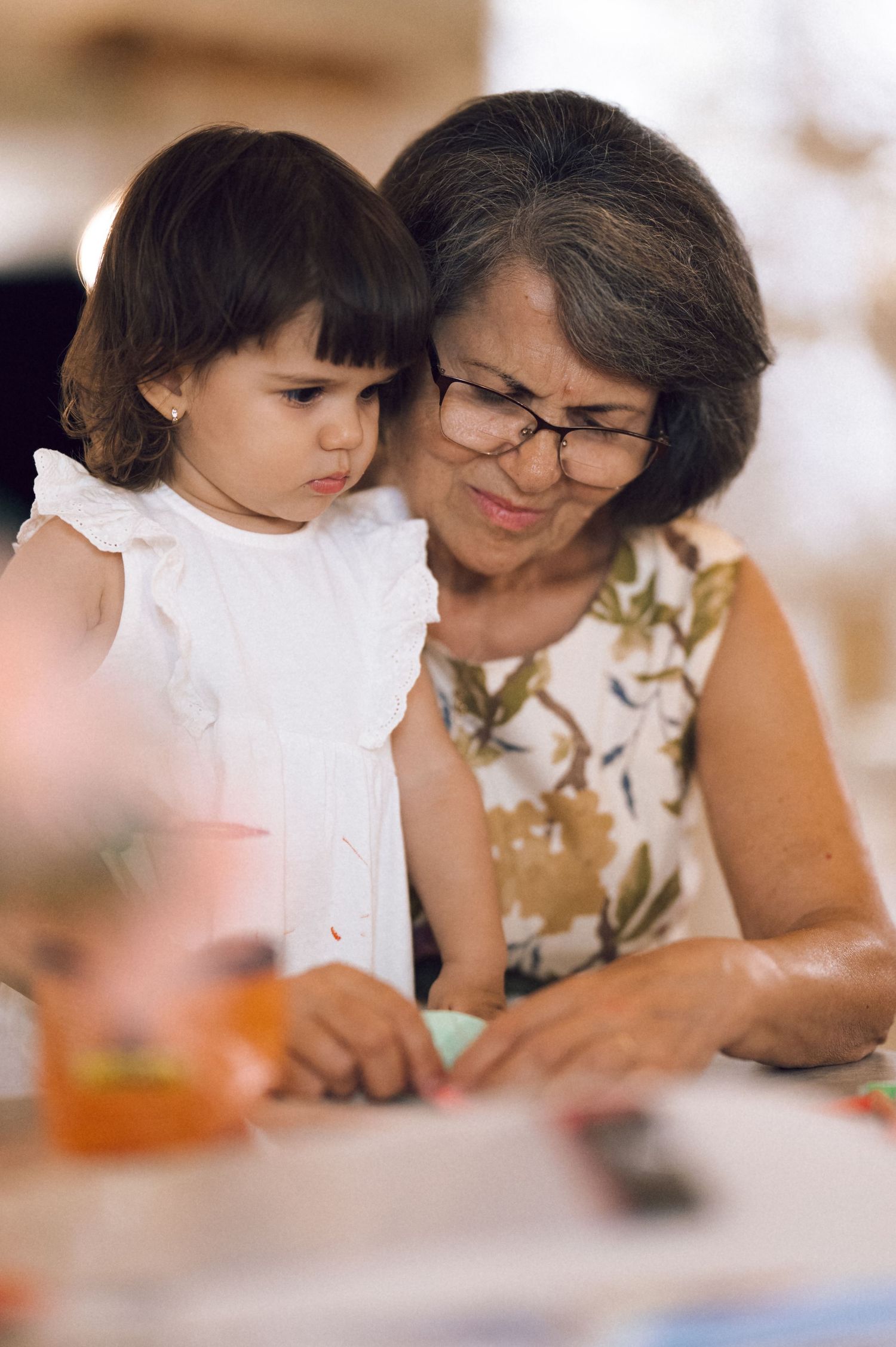 Grandmother with glasses and young toddler girl in white dress looking at something together at a table.