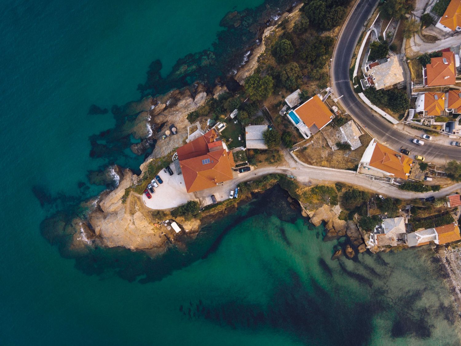 Aerial drone view of a rocky coastal peninsula with colorful houses, turquoise sea water, and a winding road.