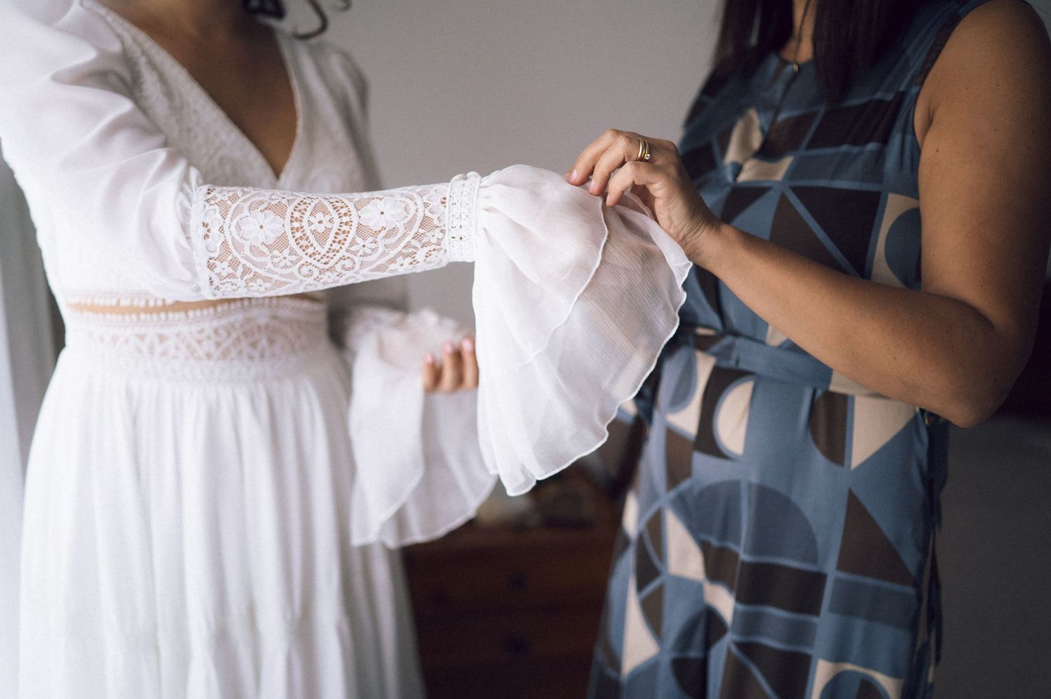 Woman in white lace wedding dress being helped by bridesmaid in blue patterned dress during bridal preparation.