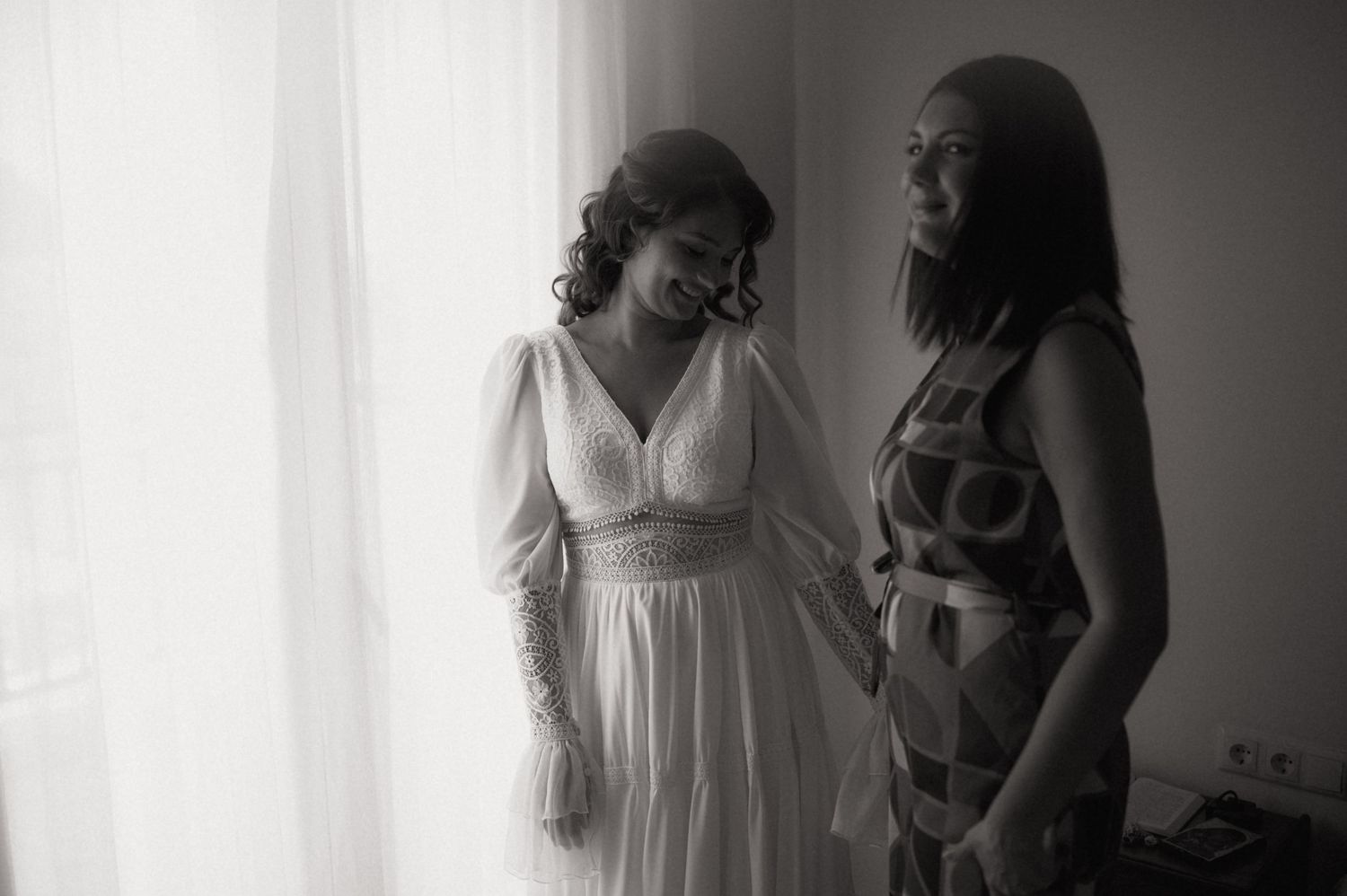 Black and white photo of a bride in white gown sharing a tender moment with a bridesmaid near a window.