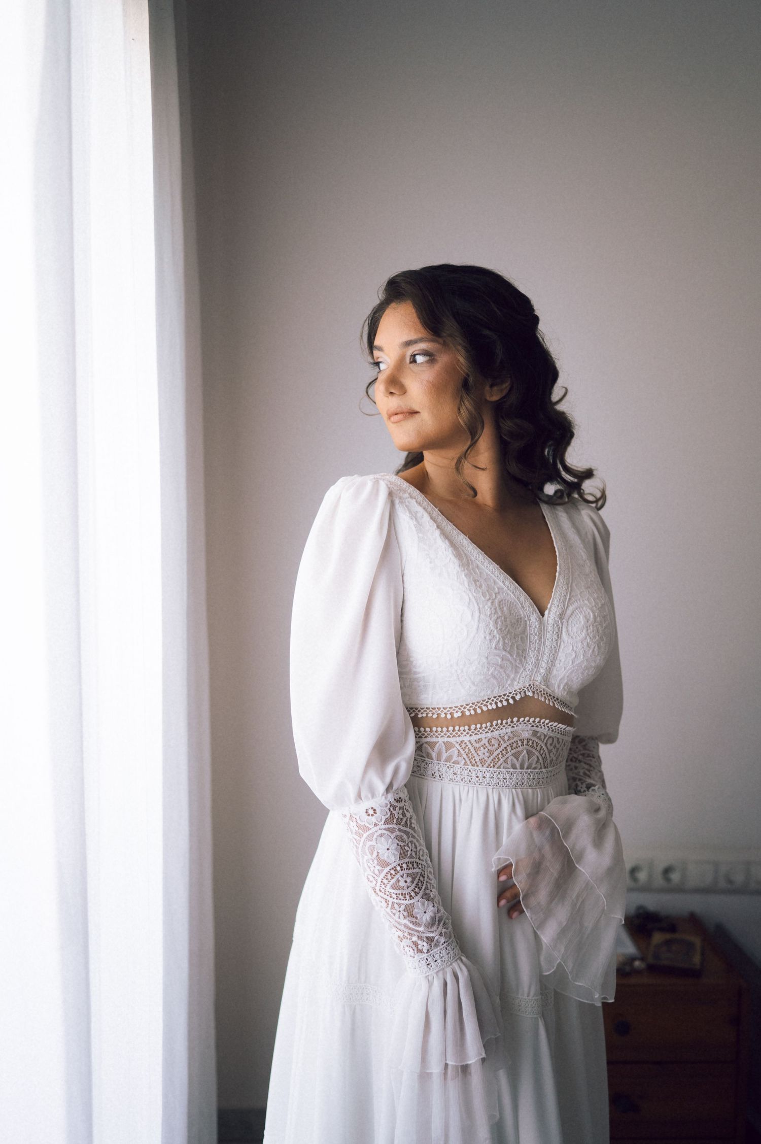 Elegant bride in white lace boho wedding dress with puff sleeves standing by window in soft natural light.