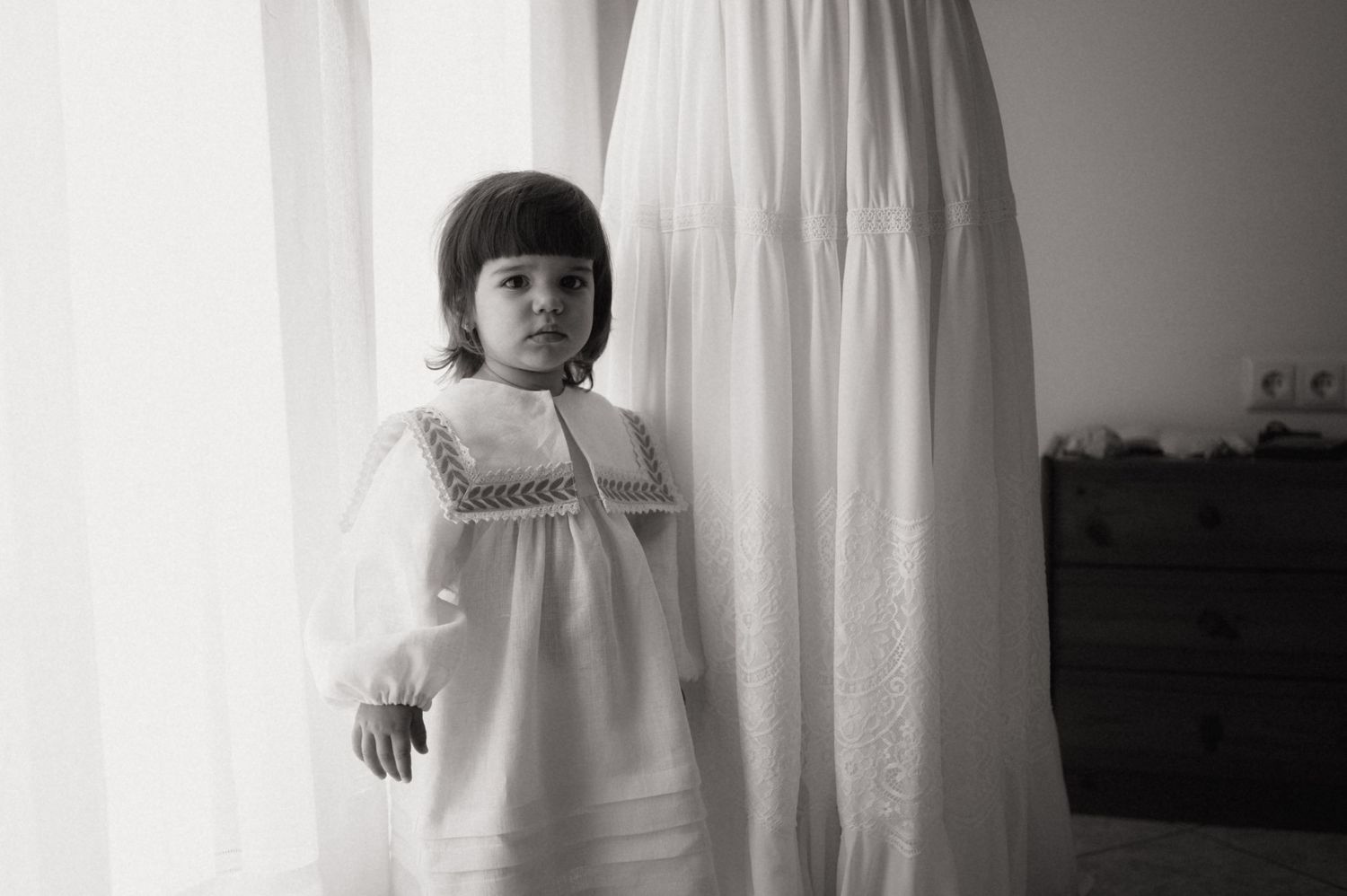 Black and white photo of a young girl in a white smocked dress standing by sheer curtains near a window.