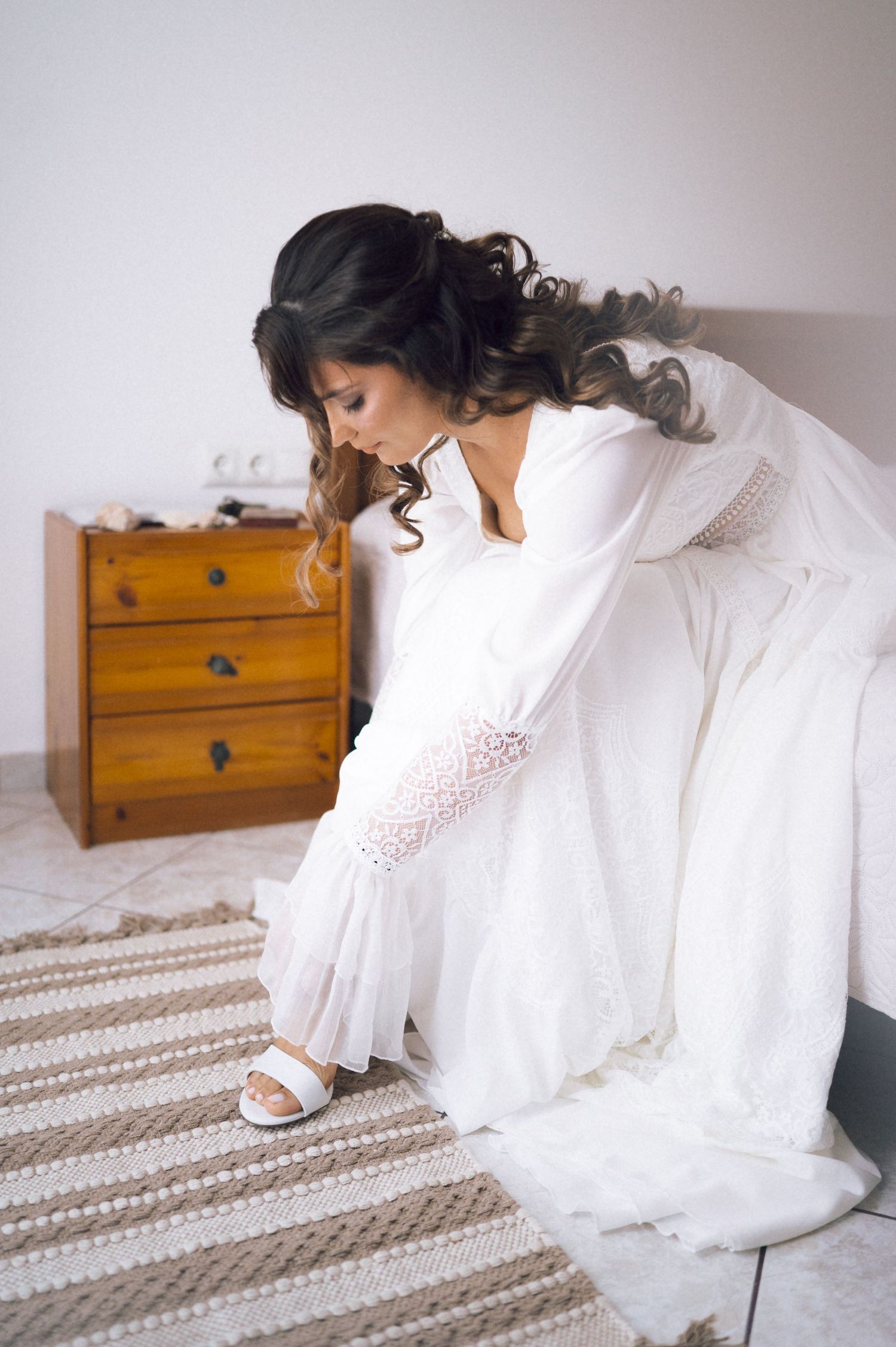 Bride in white wedding dress and lace sleeves sitting on bed putting on white sandals in bright bedroom.