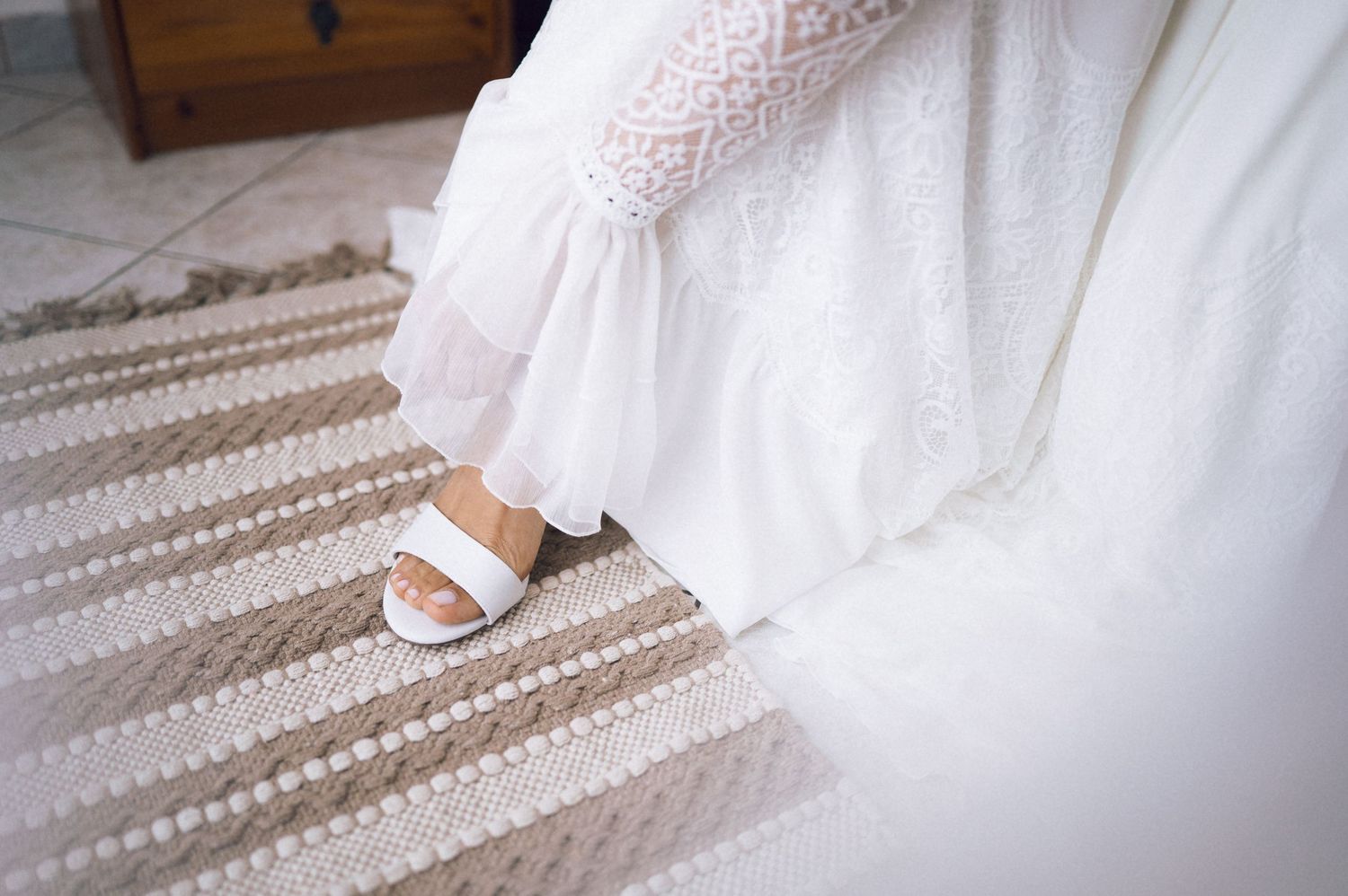 Bride wearing white lace wedding dress and white sandals standing on a striped beige rug.