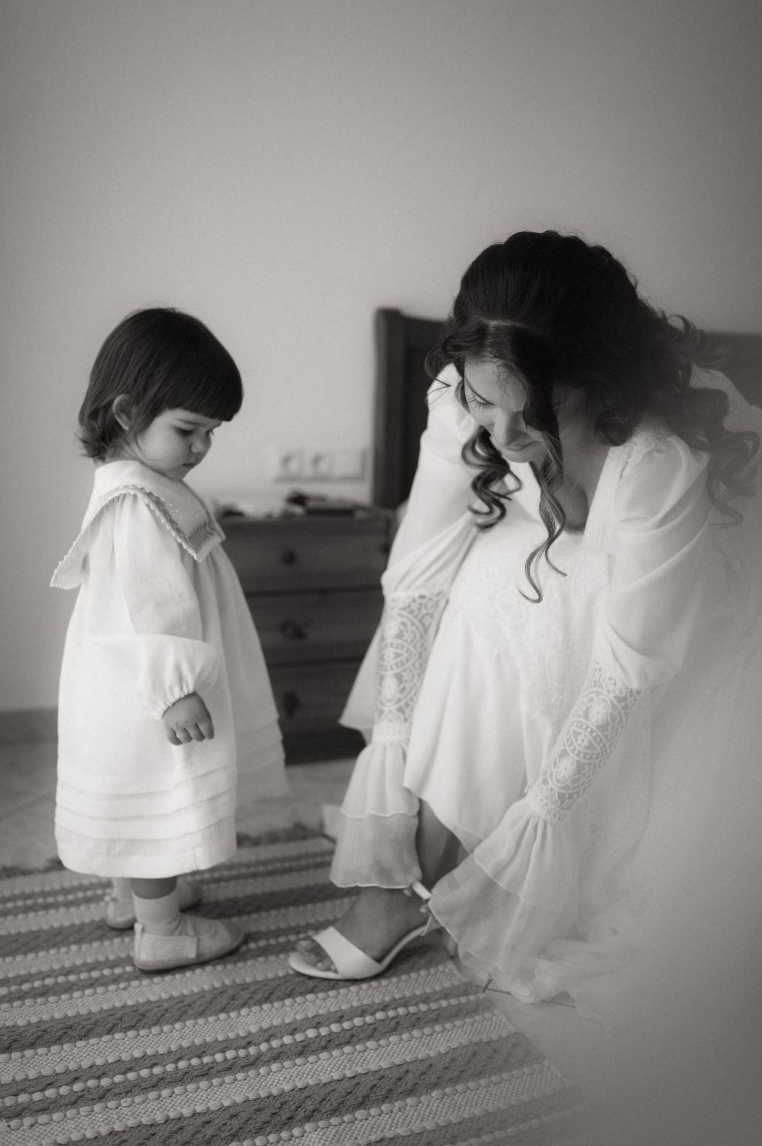 Black and white photo of a bride in white gown helping a toddler girl in white dress with shoes in a bedroom.