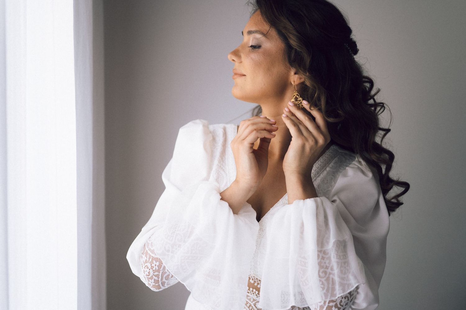 A bride in a white lace robe puts on gold earrings by a bright window, preparing for her wedding day.