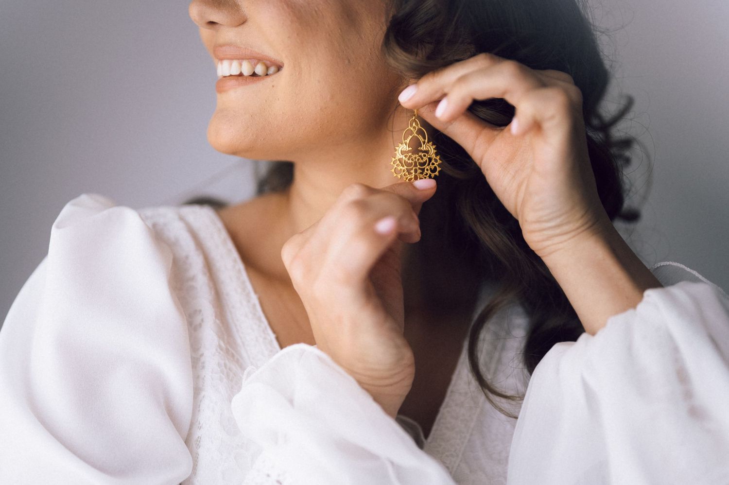 Woman smiling while putting on a gold ornate earring, wearing a white blouse against a gray background.