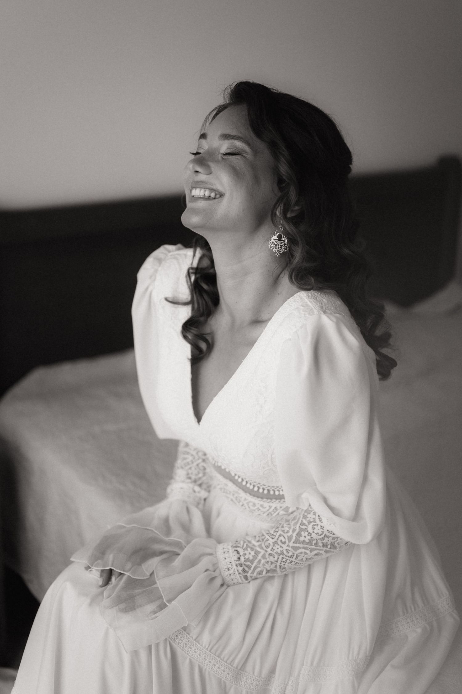Black and white photo of a smiling bride in a white lace dress sitting on a bed, laughing joyfully.