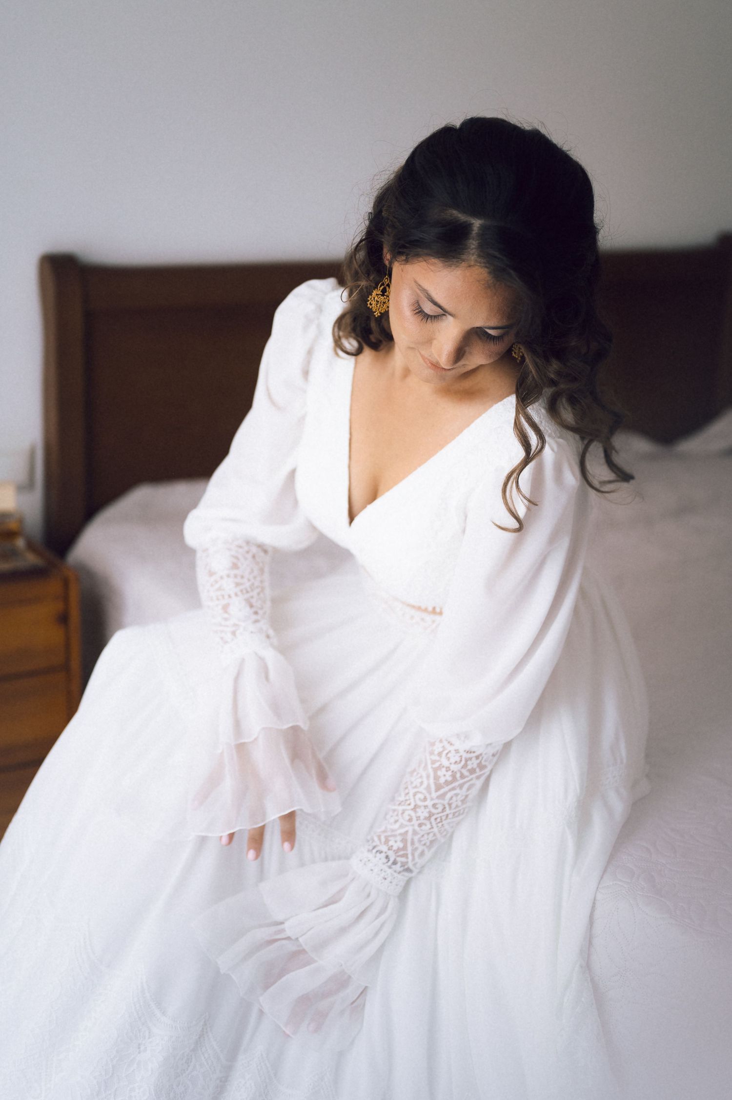 Bride in white long-sleeve lace wedding dress sitting on bed, looking down elegantly.