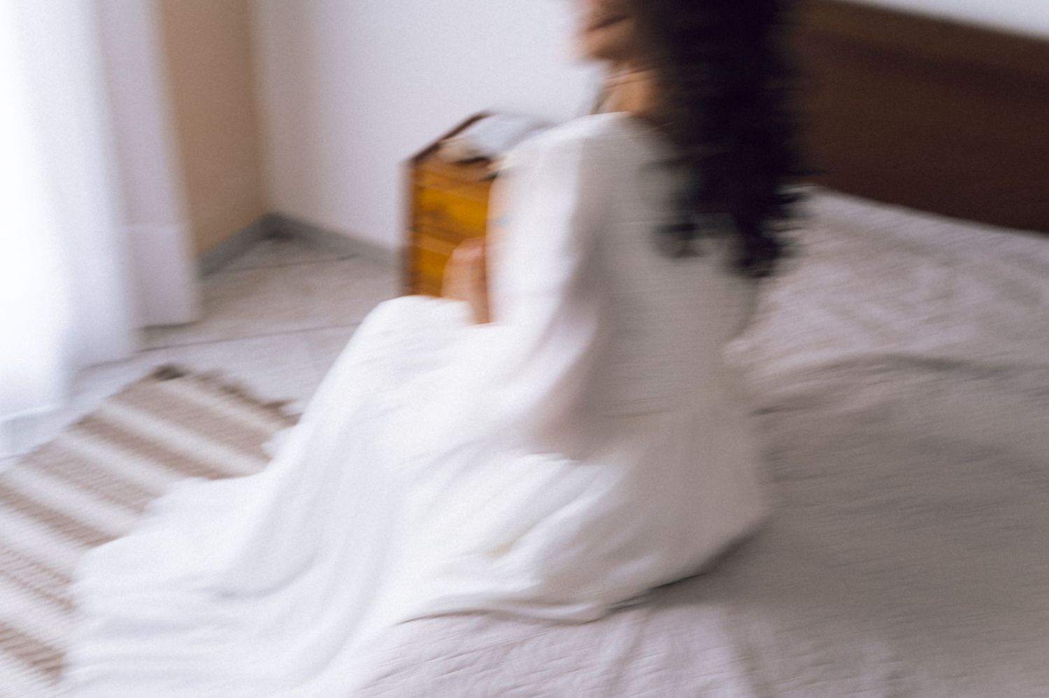 Blurred motion photo of a woman in white dress sitting on a bed holding a wooden box in a bright room.